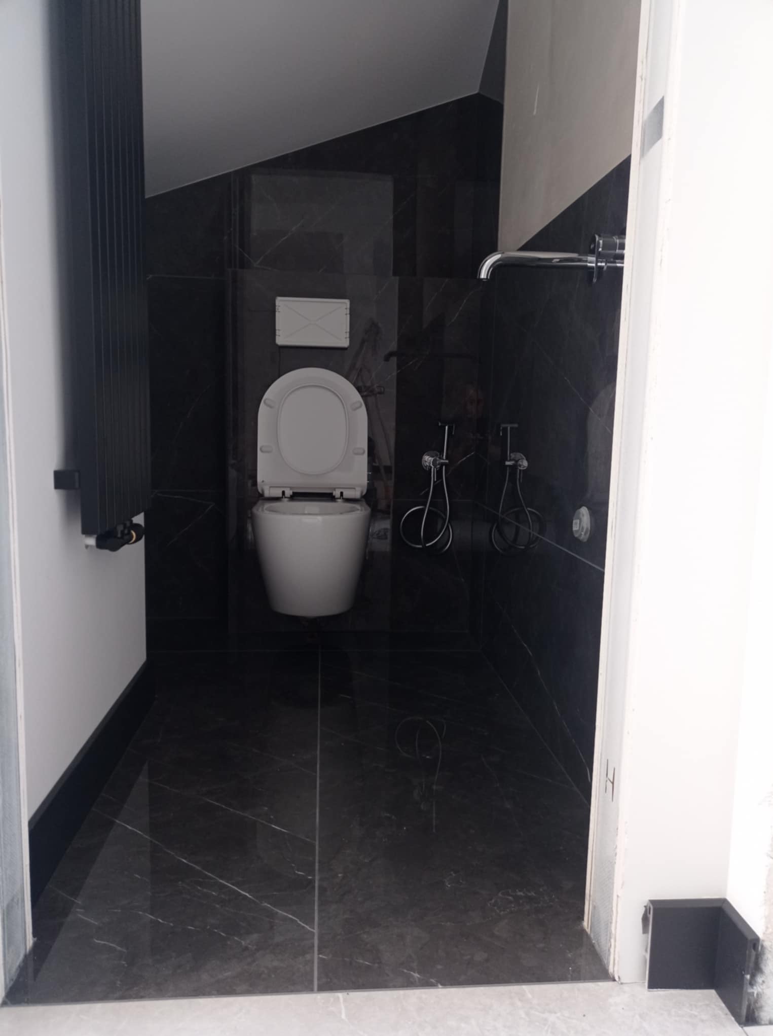 Nowoczesna łazienka z czarnymi marmurowymi płytkami, białą toaletą i chromowaną armaturą. Minimalistyczny design z otwartą przestrzenią i subtelnym oświetleniem.