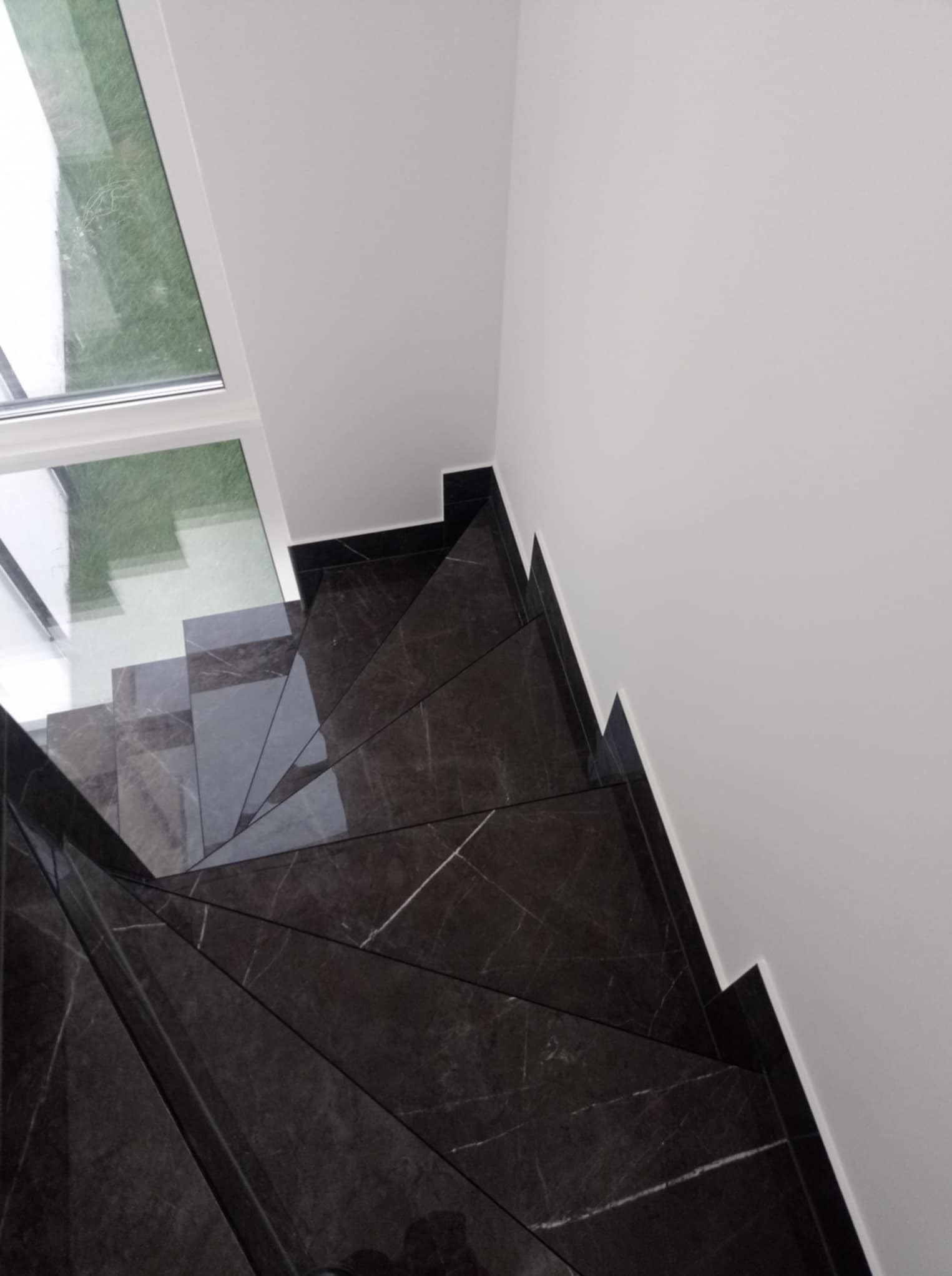 Wykończone schody z ciemnego marmuru z białymi żyłami, widok z góry, z oknem w tle. Minimalistyczne wnętrze z dbałością o detale.