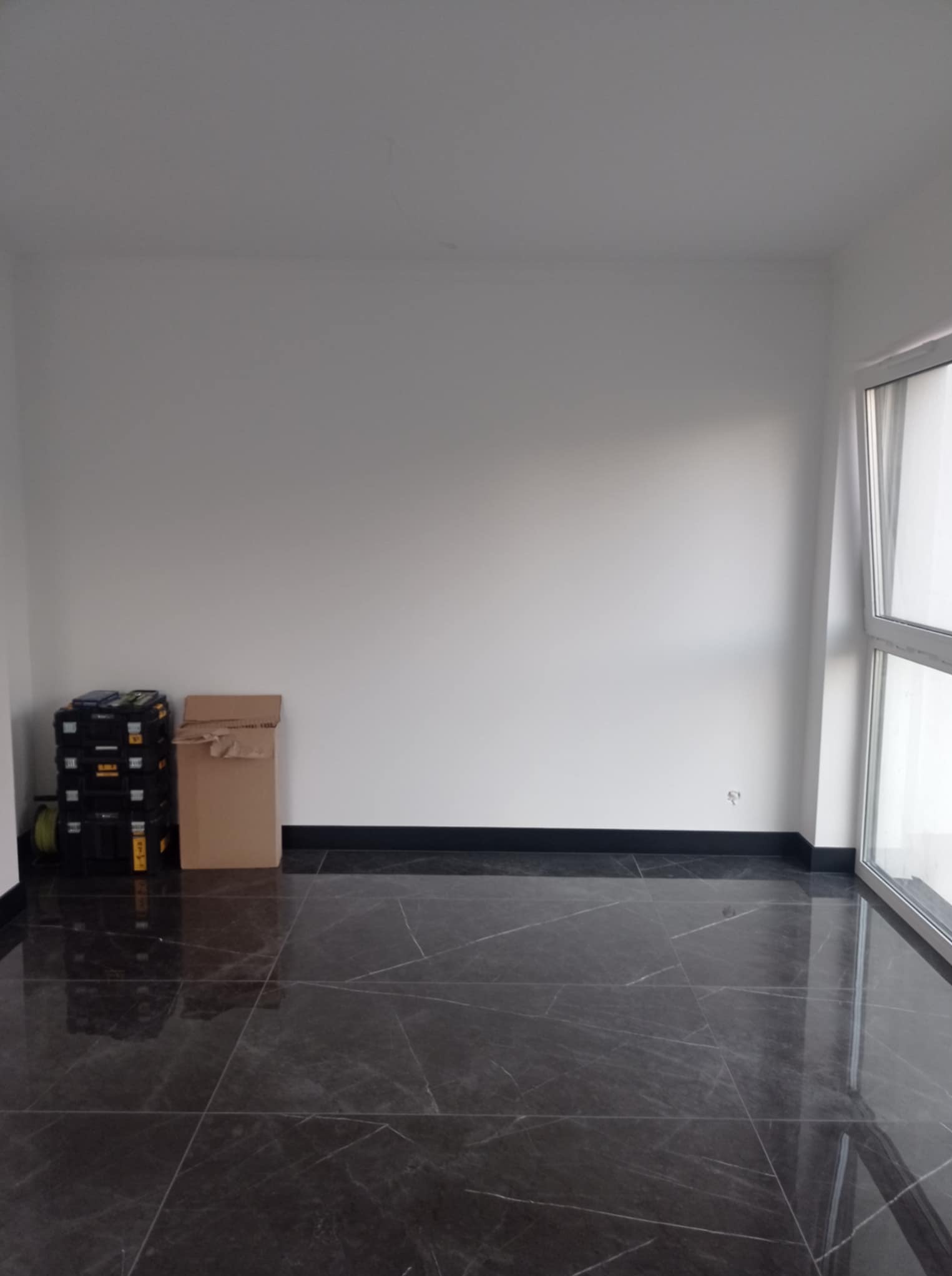 Wykończone wnętrze z ciemną, marmurkową podłogą, białe ściany i okno. W rogu narzędzia i karton. Minimalistyczny design, gotowe do umeblowania.