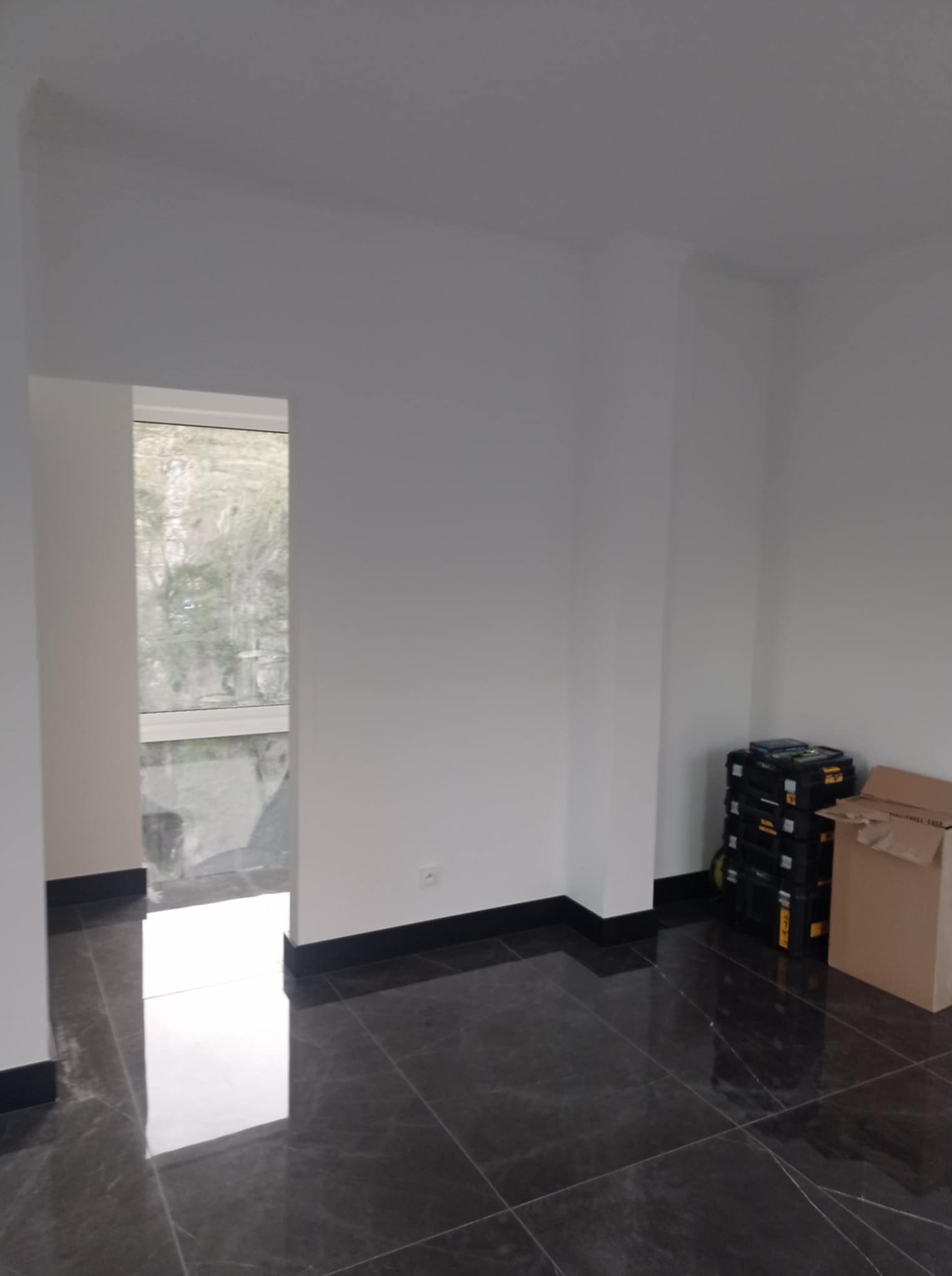 Wnętrze po remoncie: ciemna, marmurkowa podłoga z czarnymi listwami przypodłogowymi, białe ściany, okno w tle i skrzynki narzędziowe w rogu.