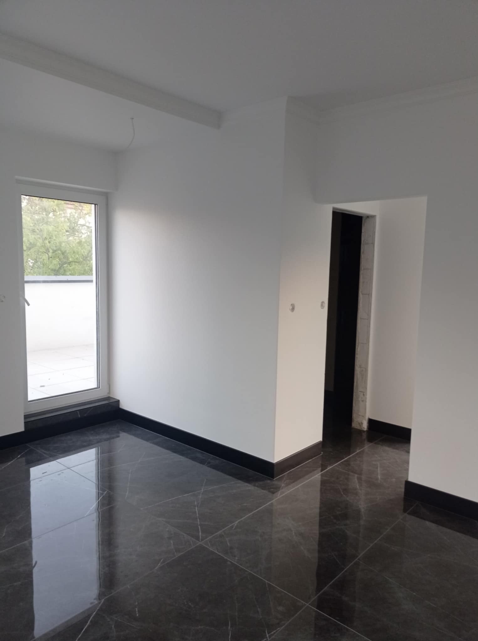 Wykończone wnętrze pokoju z ciemną, marmurkową podłogą, świeżo pomalowanymi białymi ścianami i widocznym oknem balkonowym. Minimalistyczny design.