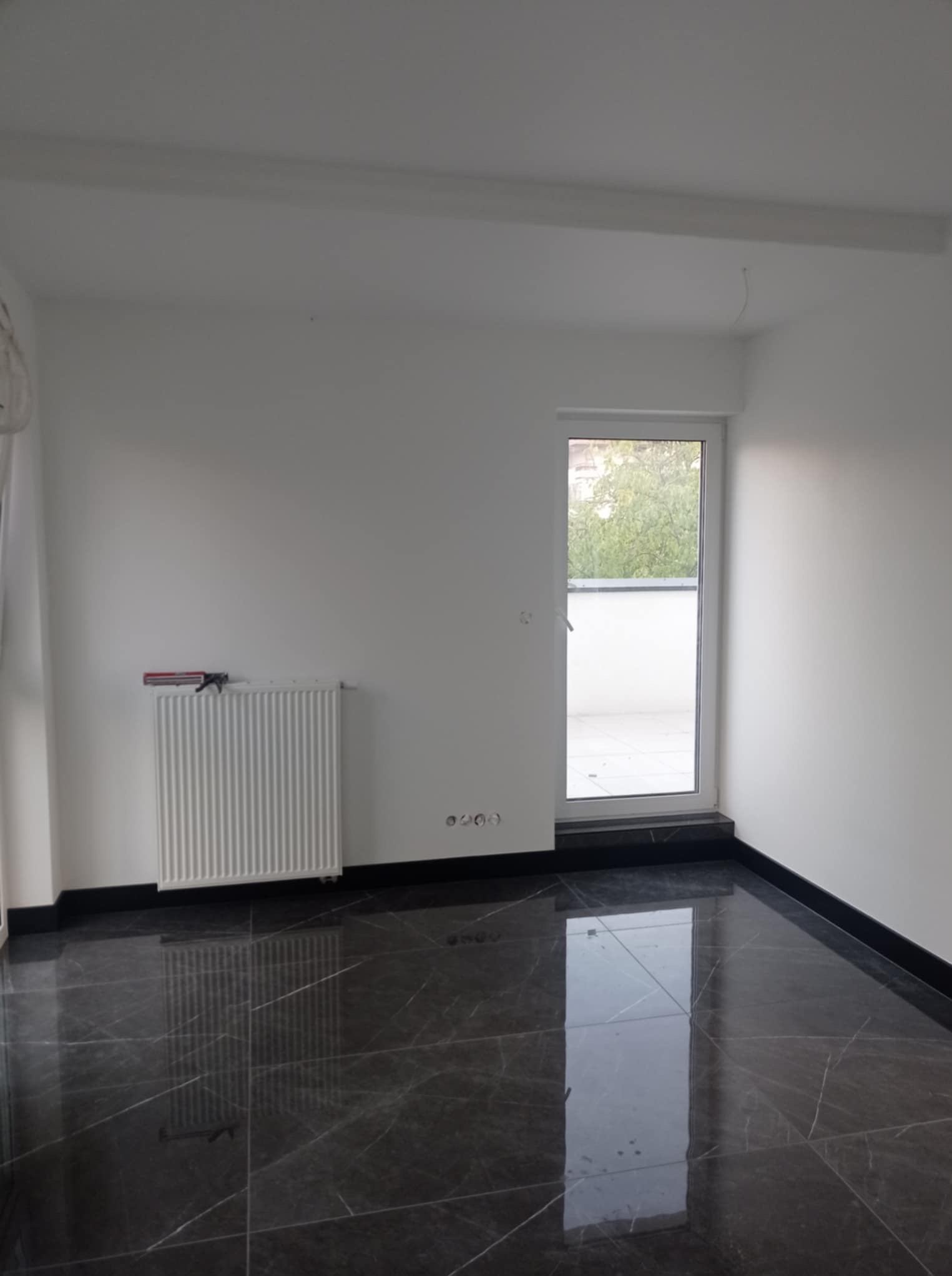 Wykończone wnętrze pokoju w Ożarowie Mazowieckim z czarną, marmurkową podłogą, białymi ścianami i grzejnikiem. Widok na balkon przez przeszklone drzwi.