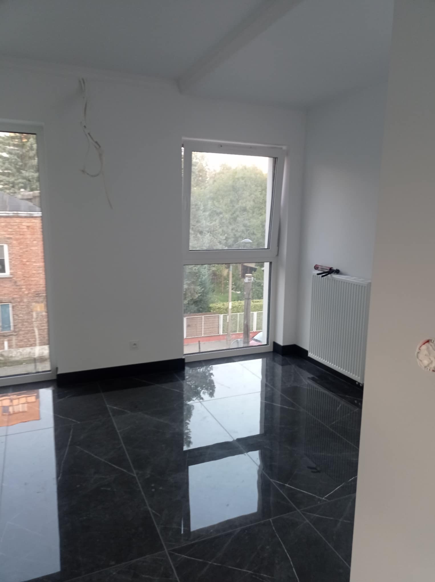 Białe ściany i lśniąca, czarna podłoga z marmuru w pustym pokoju z grzejnikiem i dwoma oknami. Widoczne kable zwisające ze ściany.