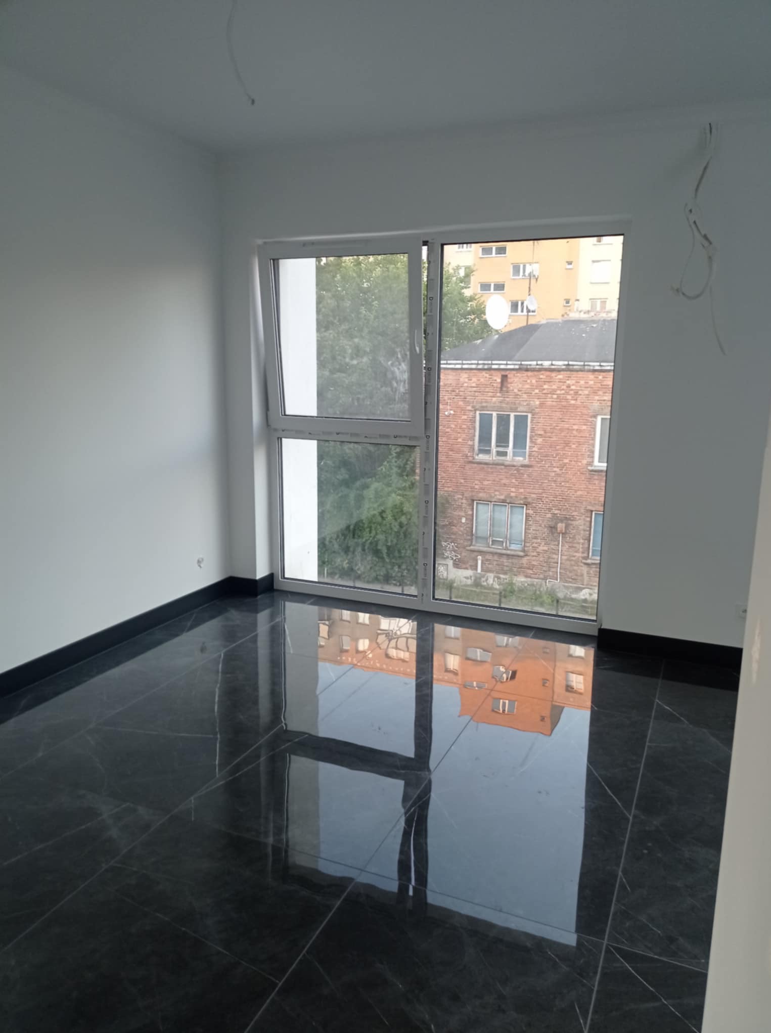 Białe ściany i błyszcząca, ciemna podłoga z marmuru w pustym pokoju po remoncie. Widok z okna na budynek z czerwonej cegły. Wiszące kable elektryczne.