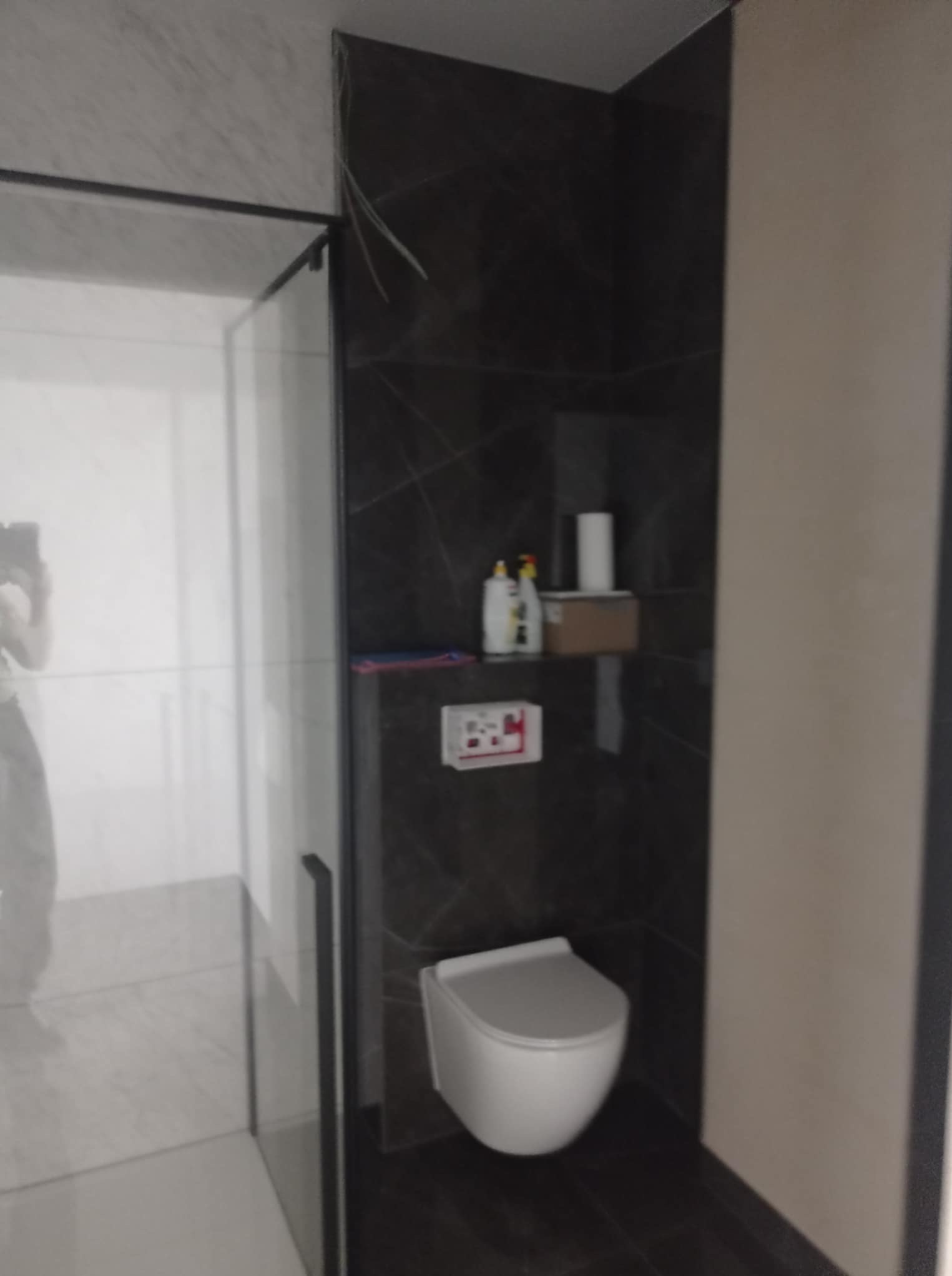 Nowoczesna łazienka po remoncie: kabina prysznicowa ze szklanymi drzwiami, wisząca toaleta i ciemne płytki imitujące marmur. Minimalistyczny design z akcentami w postaci półki.