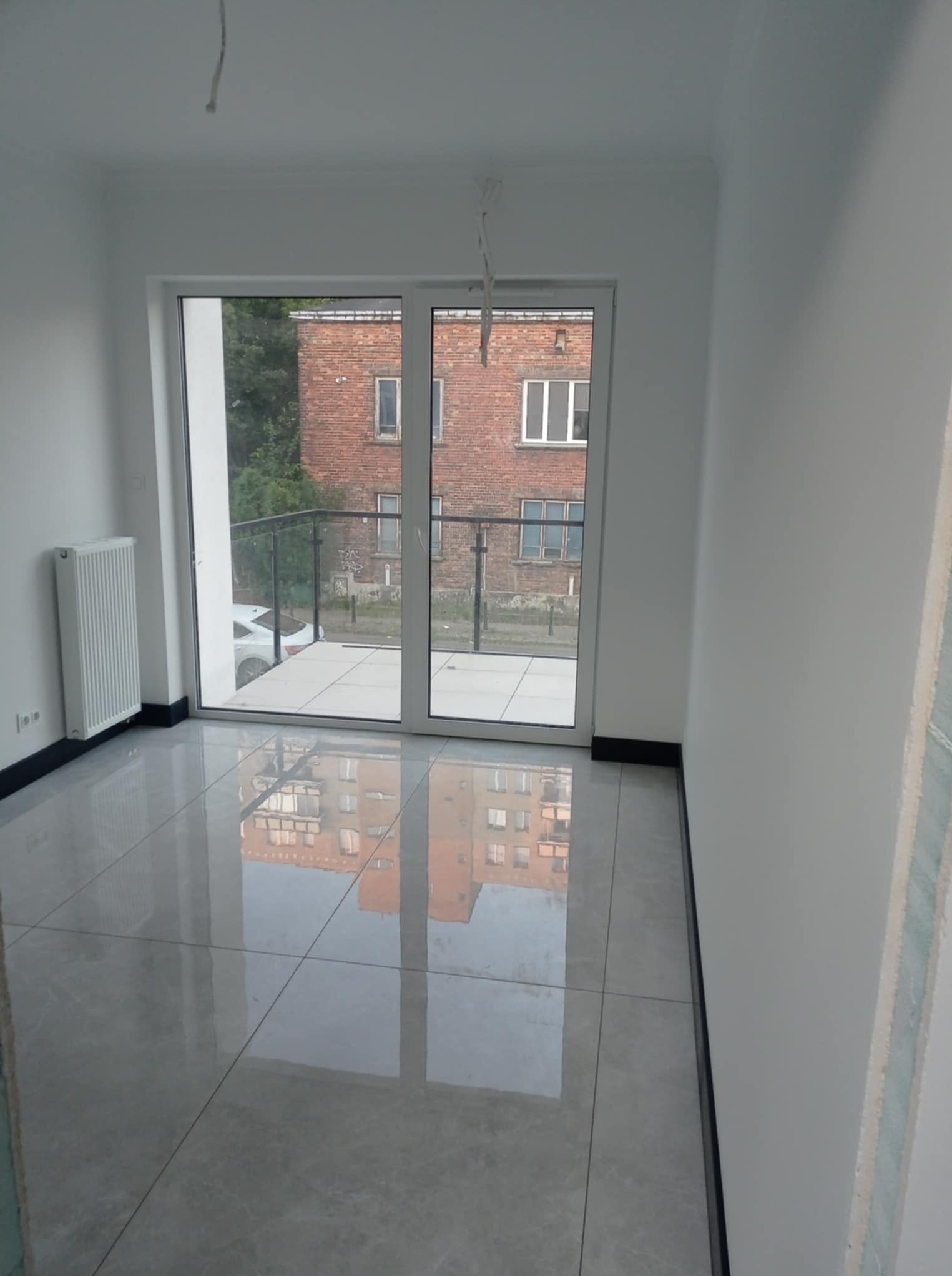 Jasne, wykończone wnętrze z błyszczącą podłogą i dużym oknem balkonowym. Widoczny grzejnik i fragment ściany w trakcie prac wykończeniowych.