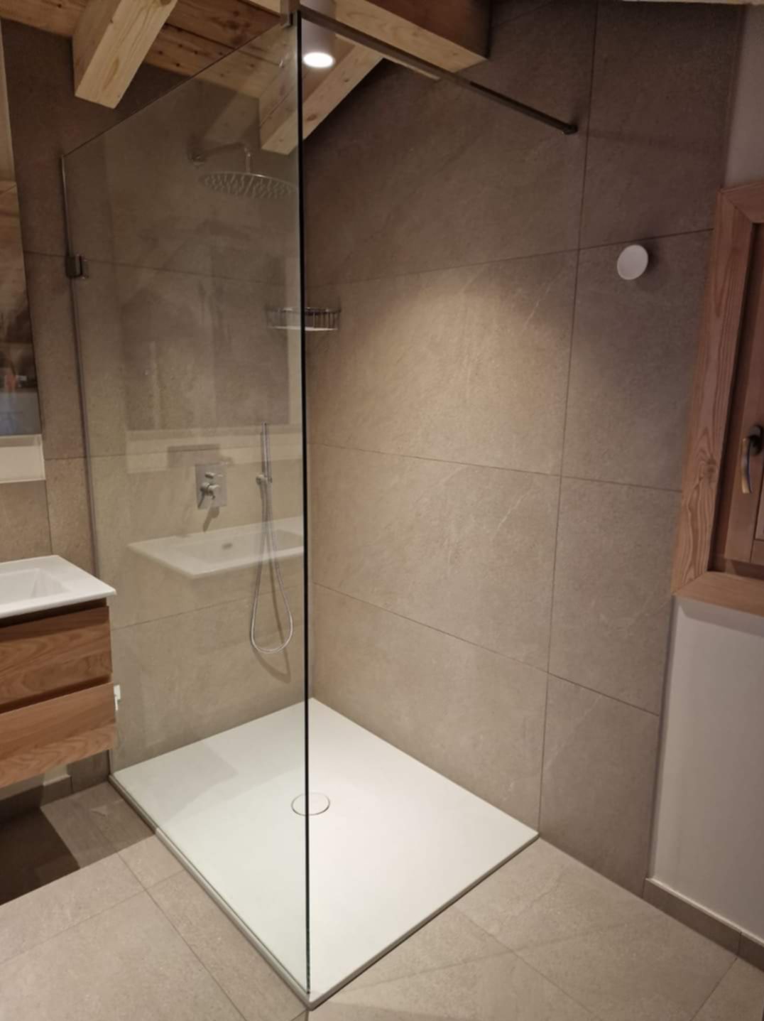 Nowoczesna kabina prysznicowa walk-in z drewnianymi elementami wykończenia. Beżowe płytki wielkoformatowe, biały brodzik i szklana ściana. Widoczna armatura i drewniana szafka z umywalką.