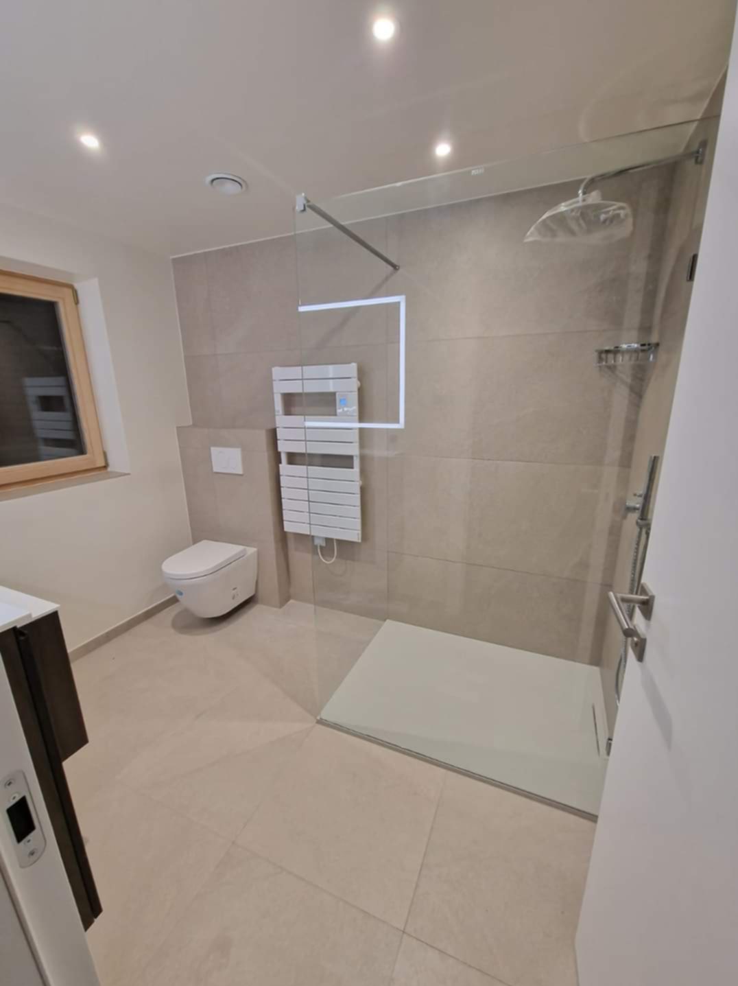 Nowoczesna łazienka z kabiną typu walk-in, beżowe płytki, podwieszana toaleta i grzejnik drabinkowy z podświetleniem LED. Minimalistyczny design, jasne wnętrze.