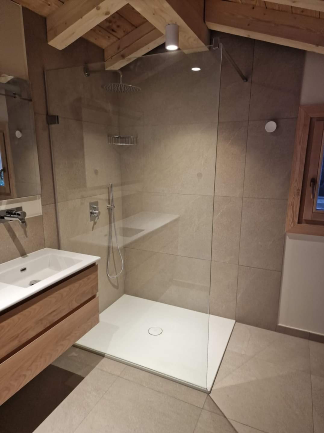 Nowoczesna łazienka z kabiną prysznicową typu walk-in, drewnianą szafką pod umywalką i beżowymi płytkami. Widoczny fragment okna z drewnianą ramą i drewniany strop.