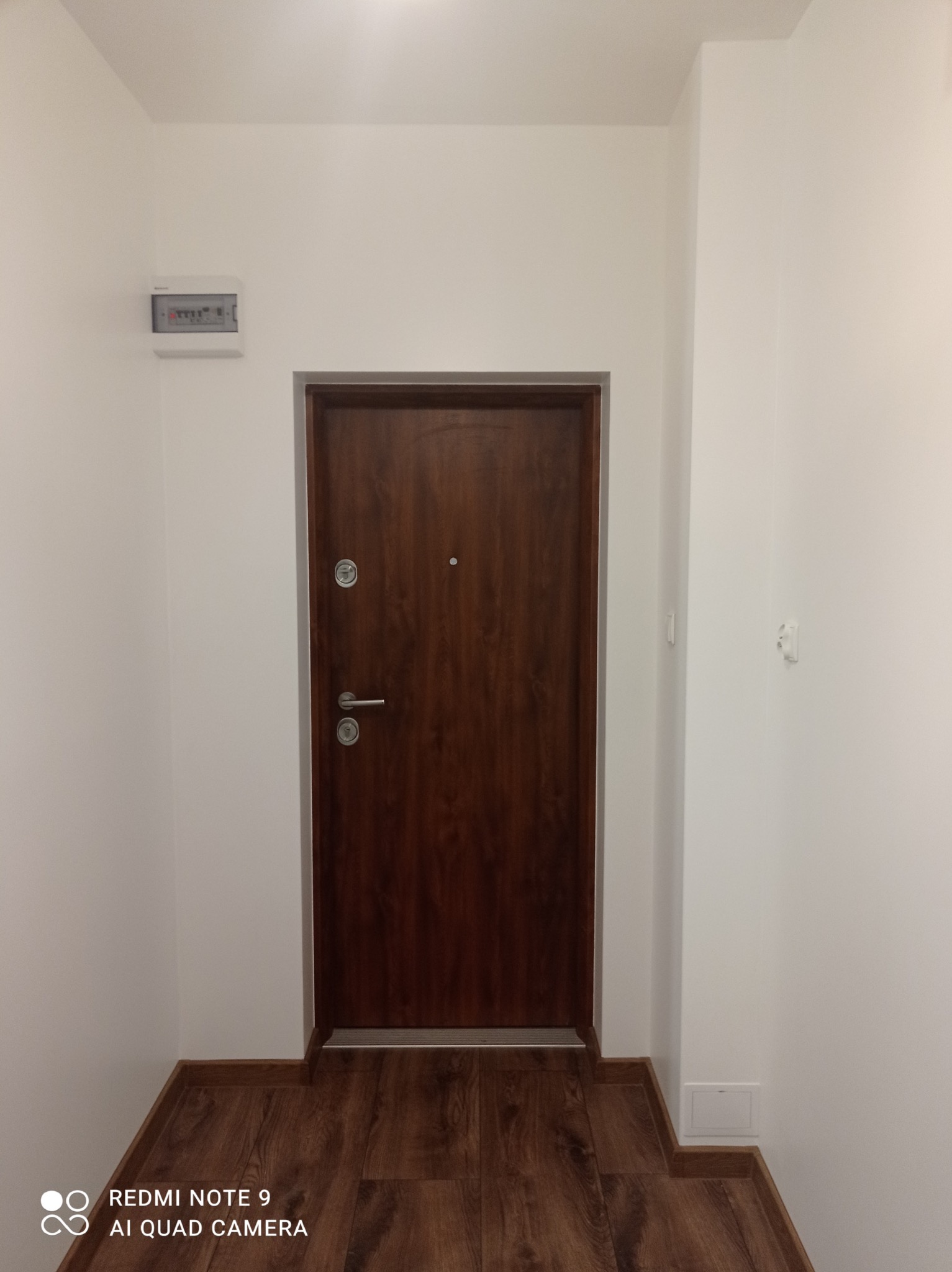 Wnętrze po remoncie: brązowe drzwi wejściowe z srebrną klamką na tle białej ściany i drewnopodobnej podłogi. Widoczna skrzynka elektryczna i listwy przypodłogowe.