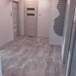 Jasne wnętrze po remoncie: drewnopodobna podłoga, białe ściany z dekoracyjnym motywem cegły, drzwi wewnętrzne z poziomymi przeszkleniami i ościeżnica w minimalistycznym stylu.