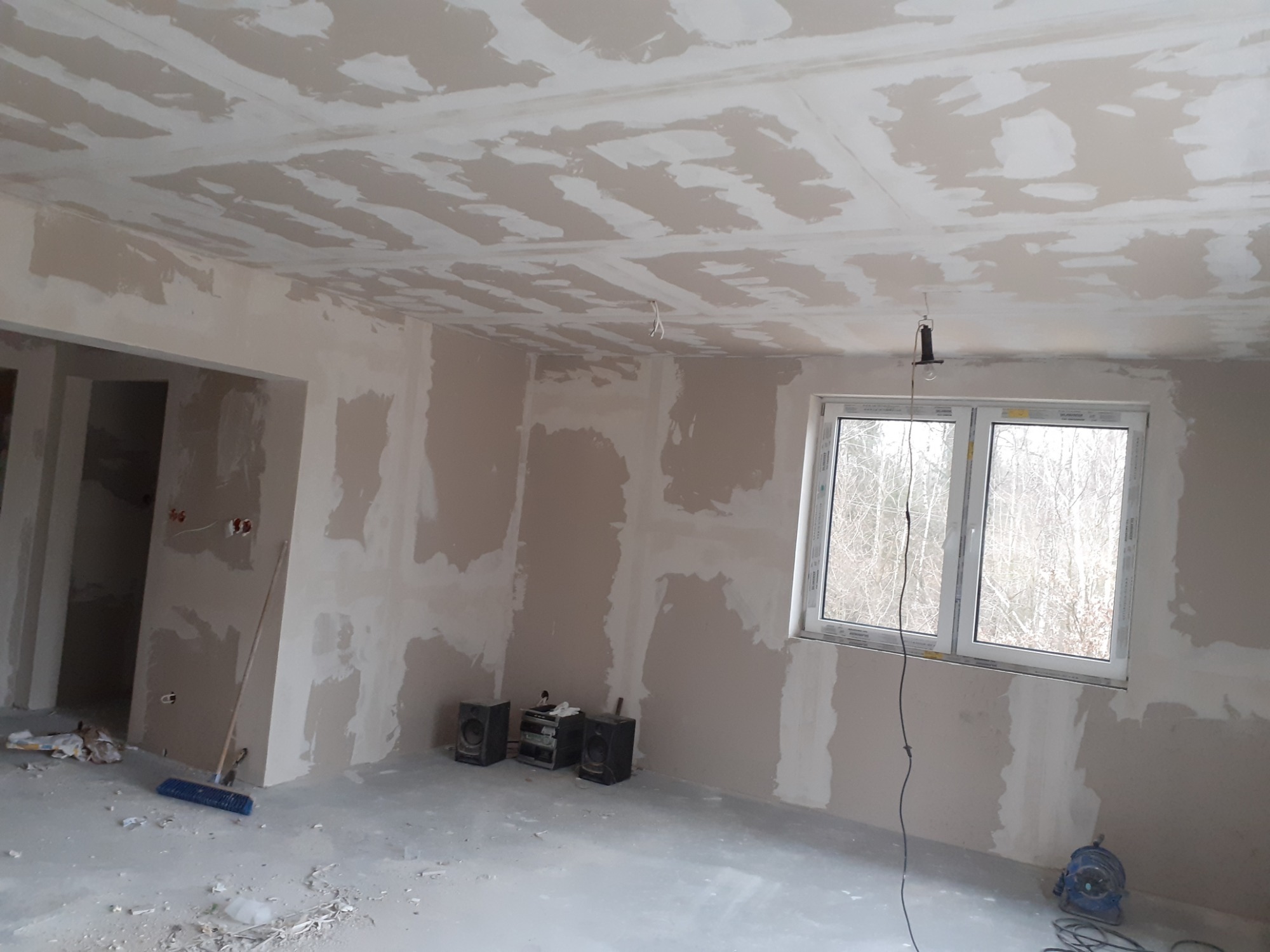 Pokój w trakcie szpachlowania: ściany i sufit pokryte warstwą gładzi, widoczne narzędzia i sprzęt audio. Okno z widokiem na drzewa. Etap prac wykończeniowych.