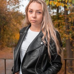 Magoń Fotografia i Film Dominik Magoń - Portret kobiety z blond włosami w czarnej skórzanej kurtce na tle jesiennego parku. Ujęcie z lekkiej góry, widoczne detale biżuterii i zegarka.