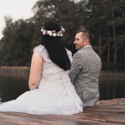 Magoń Fotografia i Film Dominik Magoń - Para młoda siedzi na drewnianym pomoście nad jeziorem. Panna młoda w sukni z koronką i wiankiem, pan młody w marynarce w kratę. Romantyczna sceneria, stonowana kolorystyka.