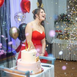 Magoń Fotografia i Film Dominik Magoń - Uśmiechnięta dziewczyna w czerwonej sukience, z opaską '18', stoi za tortem urodzinowym w otoczeniu balonów i konfetti podczas imprezy.