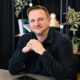 Magoń Fotografia i Film Dominik Magoń - Mężczyzna w okularach, w czarnej koszuli, siedzi za biurkiem z laptopem. W tle regał z roślinami i książkami. Uśmiechnięty, z rękami złożonymi na blacie.