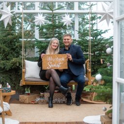 Magoń Fotografia i Film Dominik Magoń - Uśmiechnięta para siedzi na huśtawce w szklarni, trzymając tabliczkę z napisem 'Wesołych Świąt', otoczona choinkami i dekoracjami świątecznymi.