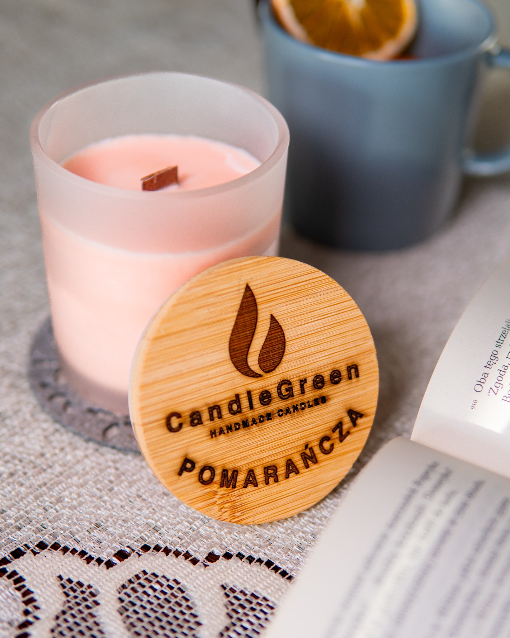 Ręcznie robiona świeca sojowa CandleGreen o zapachu pomarańczy, z drewnianym knotem, na tle książki i kubka z pomarańczą, na dekoracyjnej serwetce.