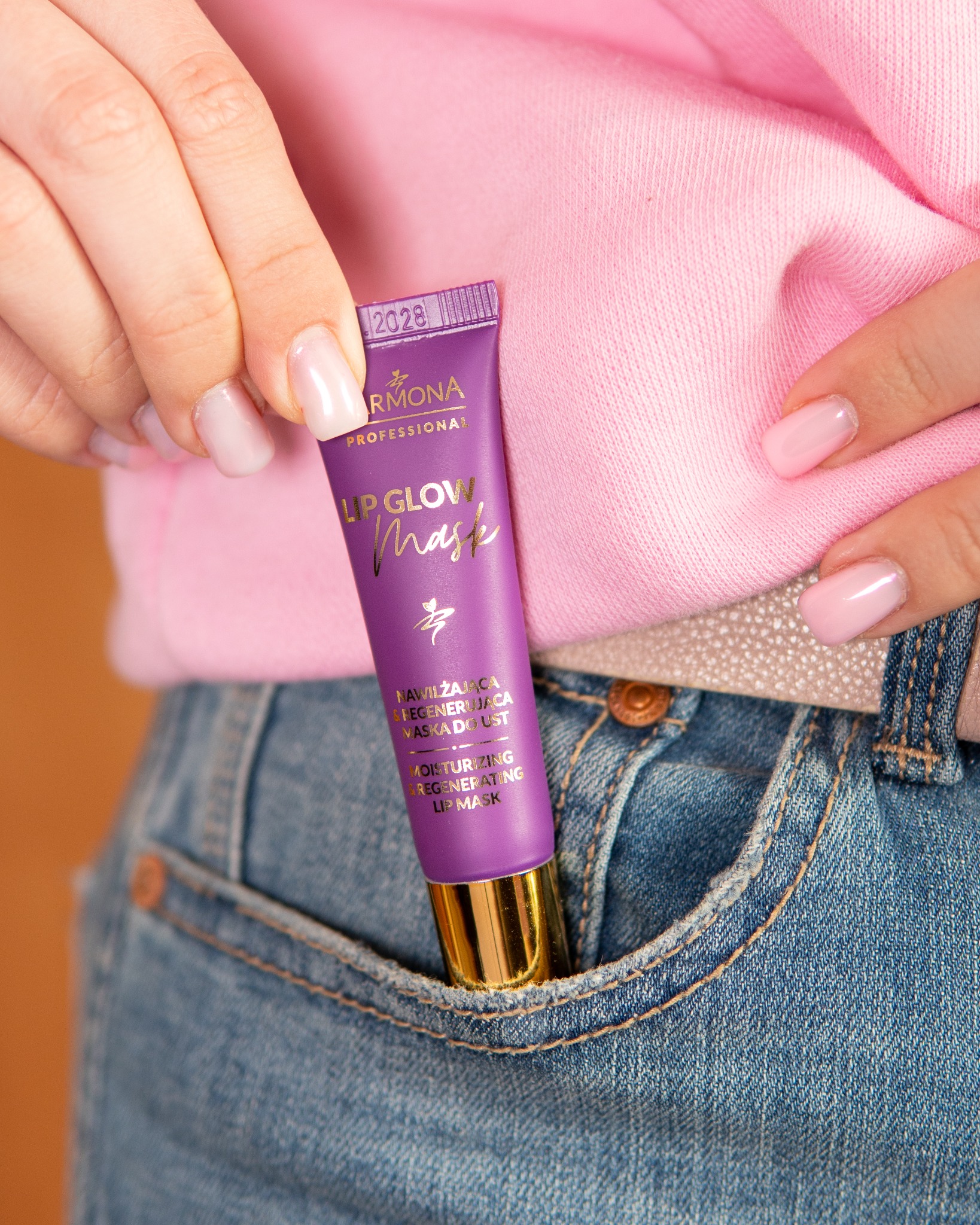 Fioletowa maseczka do ust Armona w kieszonce jeansów, trzymana przez dłoń z jasnoróżowym manicure. Ujęcie z bliska, na pierwszym planie produkt i dłoń.