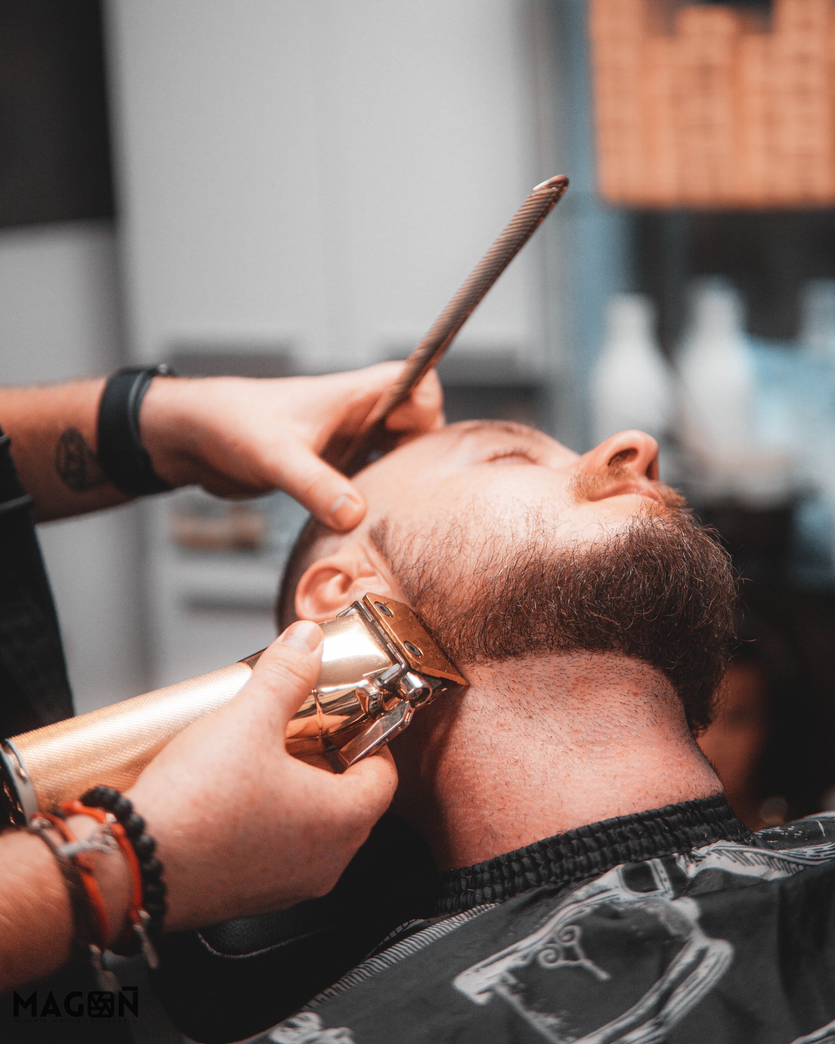 Strzyżenie brody złotym trymerem w salonie barberskim. Fryzjer z tatuażem przycina brodę klientowi leżącemu na fotelu. W tle butelki kosmetyków.