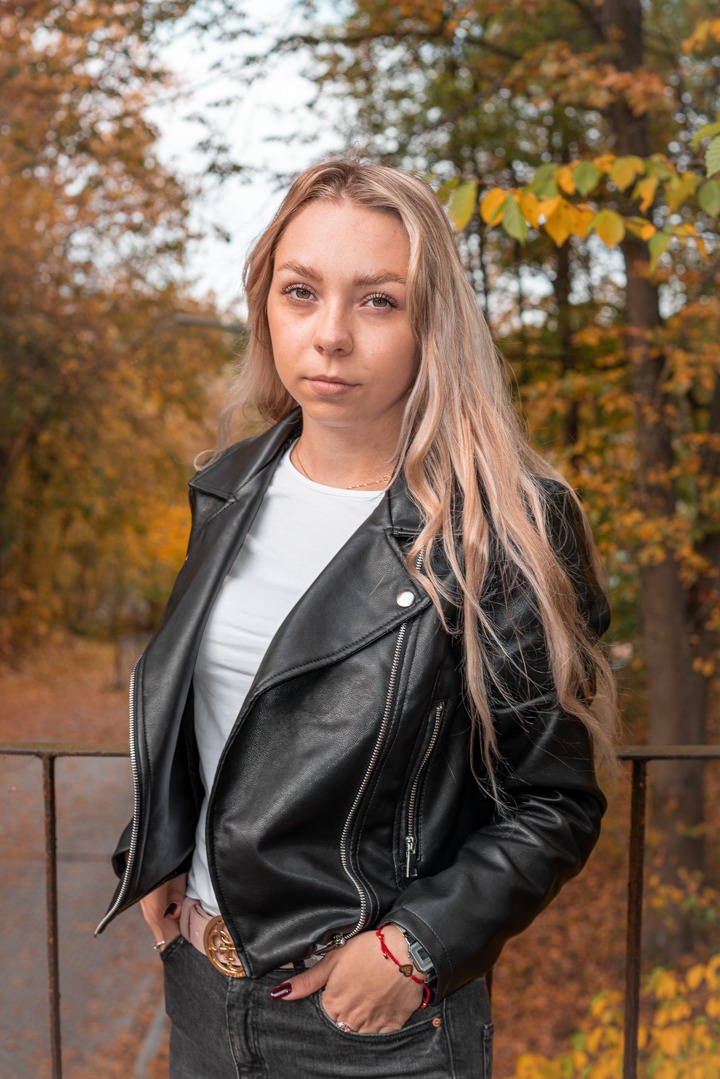 Portret kobiety z blond włosami w czarnej skórzanej kurtce na tle jesiennego parku. Ujęcie z lekkiej góry, widoczne detale biżuterii i zegarka.