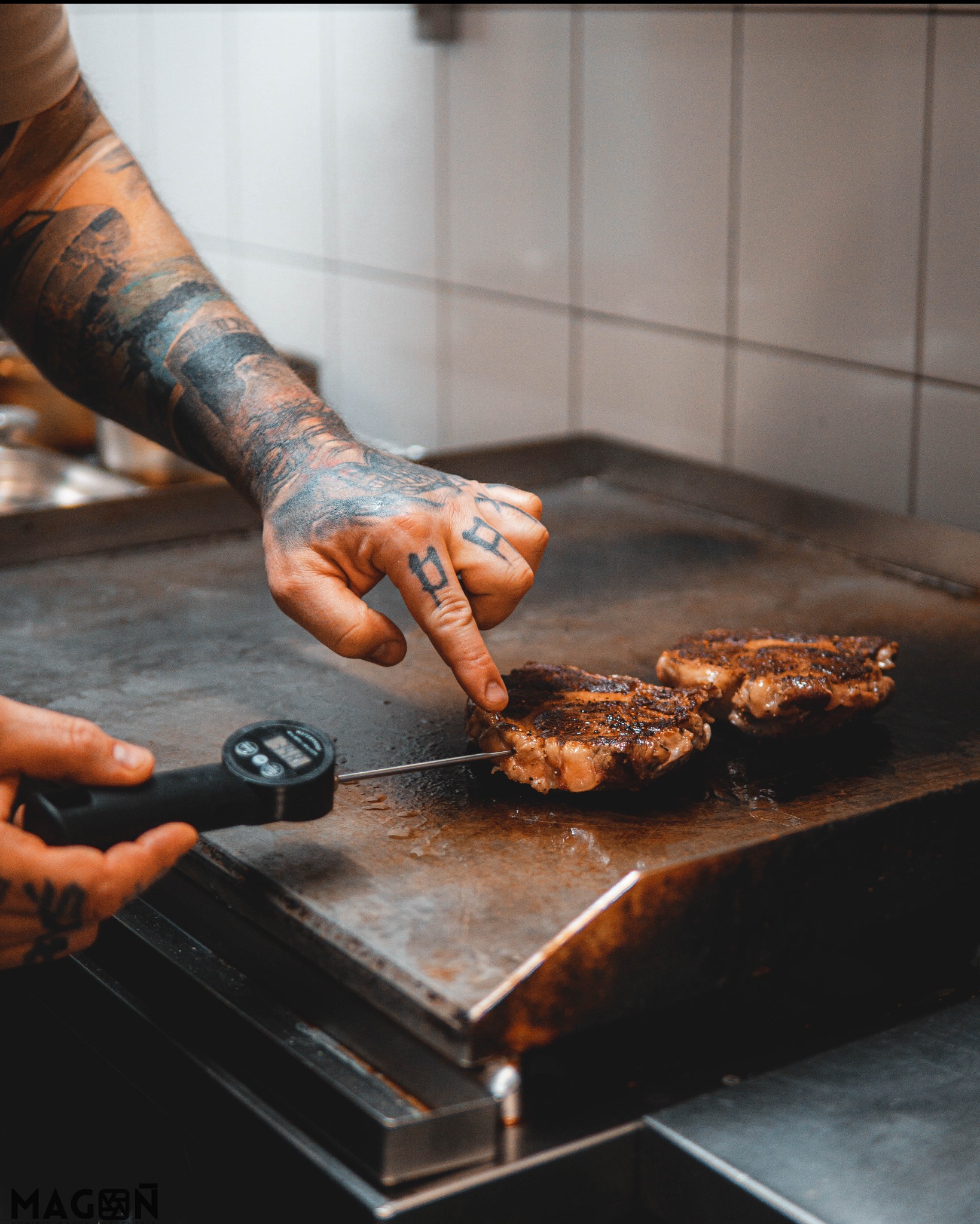 Tatuator mierzy temperaturę steków na płycie grilla. Widoczne dłonie z tatuażami i termometr. Kulinarna precyzja w akcji.