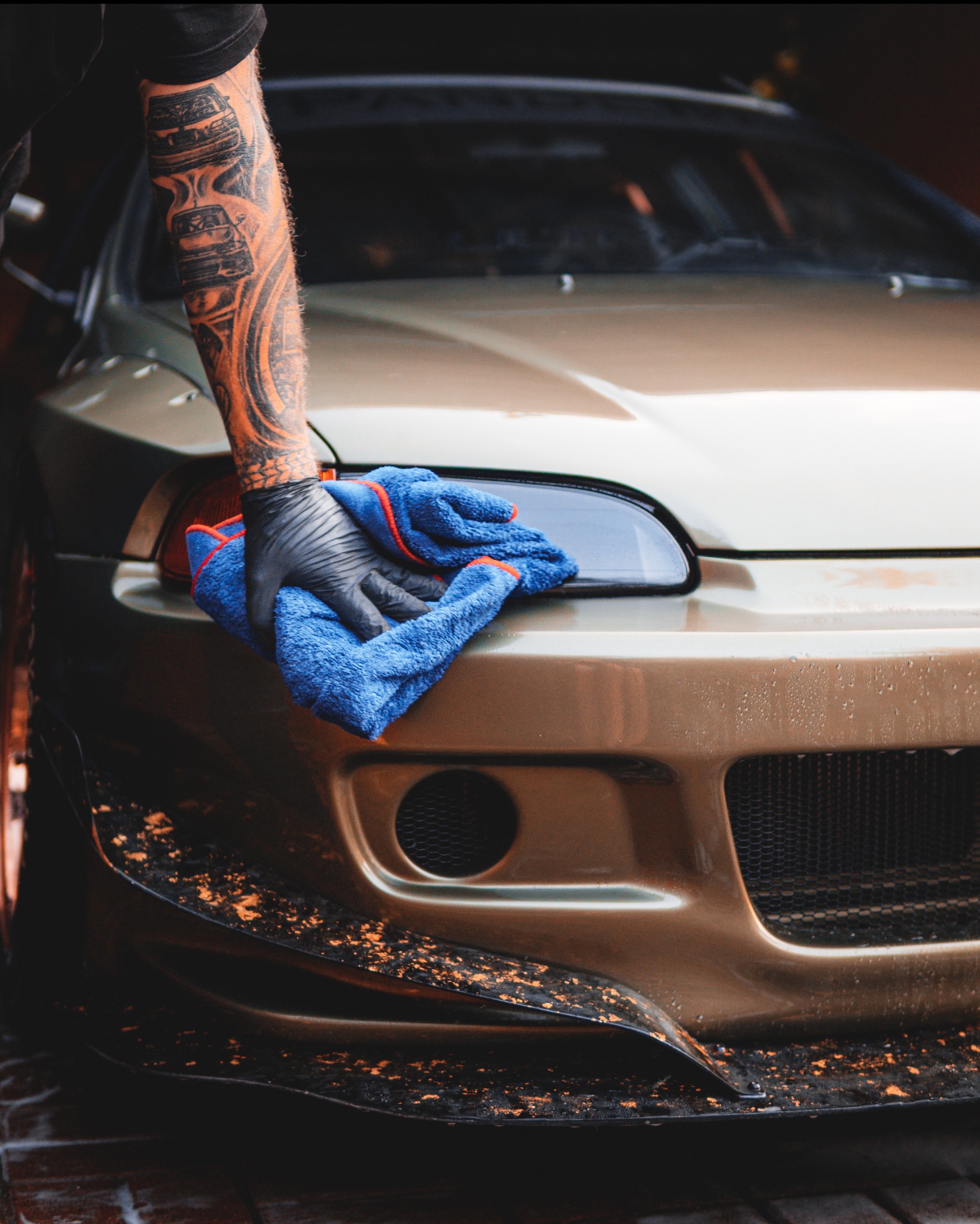 Złoty samochód czyszczony niebieską ściereczką przez osobę z tatuażami na ramieniu i w czarnych rękawicach. Detailing auta, pielęgnacja lakieru.
