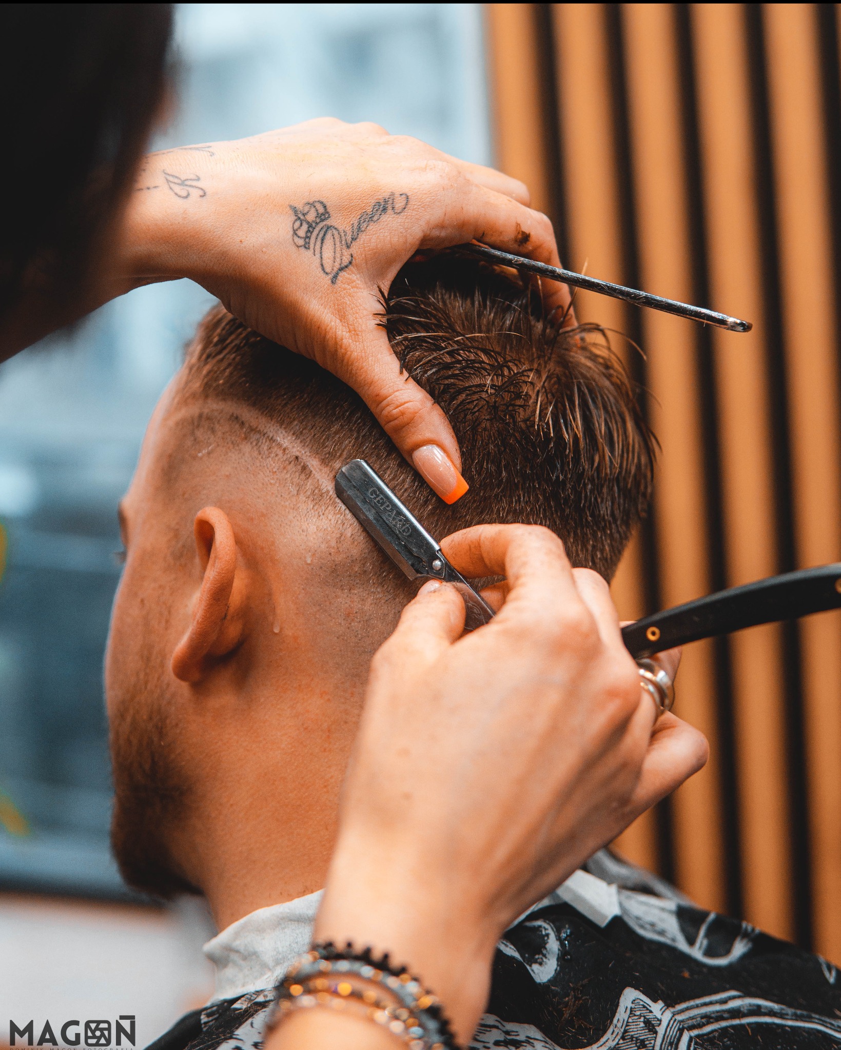 Golenie brzytwą włosów na głowie klienta w salonie barberskim. W tle pionowe, brązowe panele. Na dłoni barbera tatuaż korony i napis Queen.