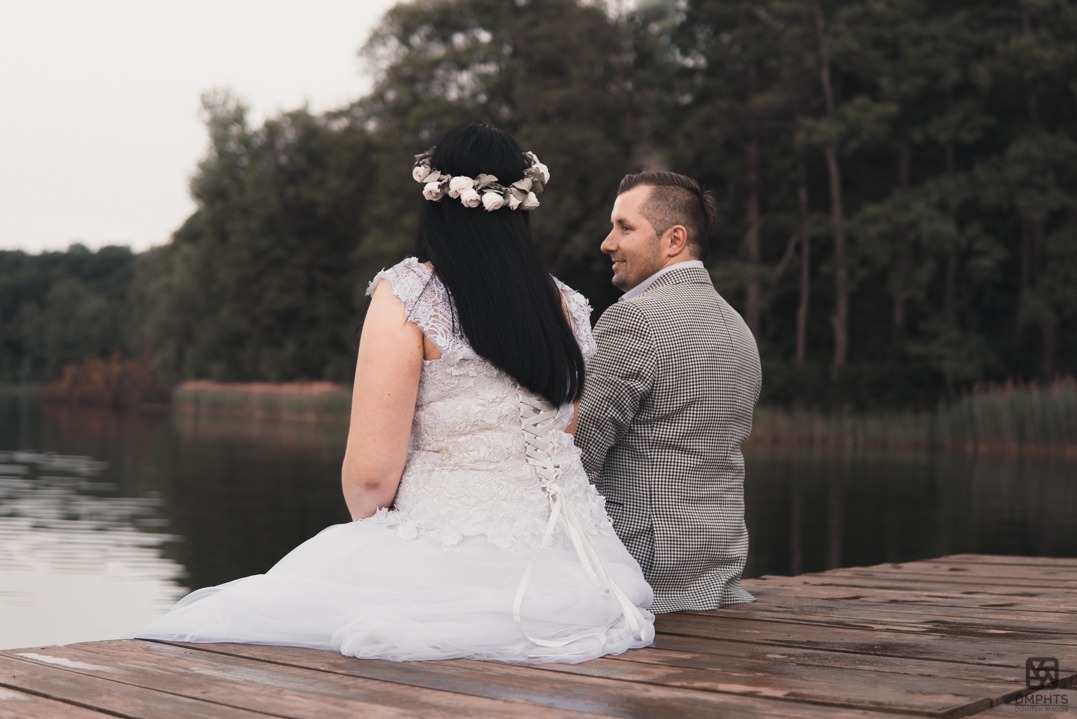Para młoda siedzi na drewnianym pomoście nad jeziorem. Panna młoda w sukni z koronką i wiankiem, pan młody w marynarce w kratę. Romantyczna sceneria, stonowana kolorystyka.