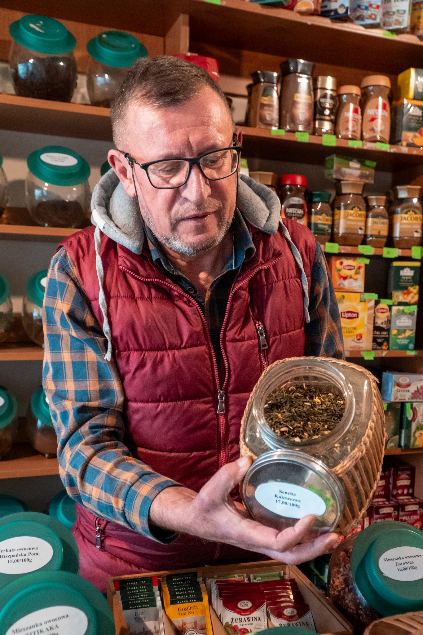 Sprzedawca prezentuje słoik z zieloną herbatą Sencha Kaktusowa w sklepie z herbatami i kawami, na tle półek z różnymi produktami.