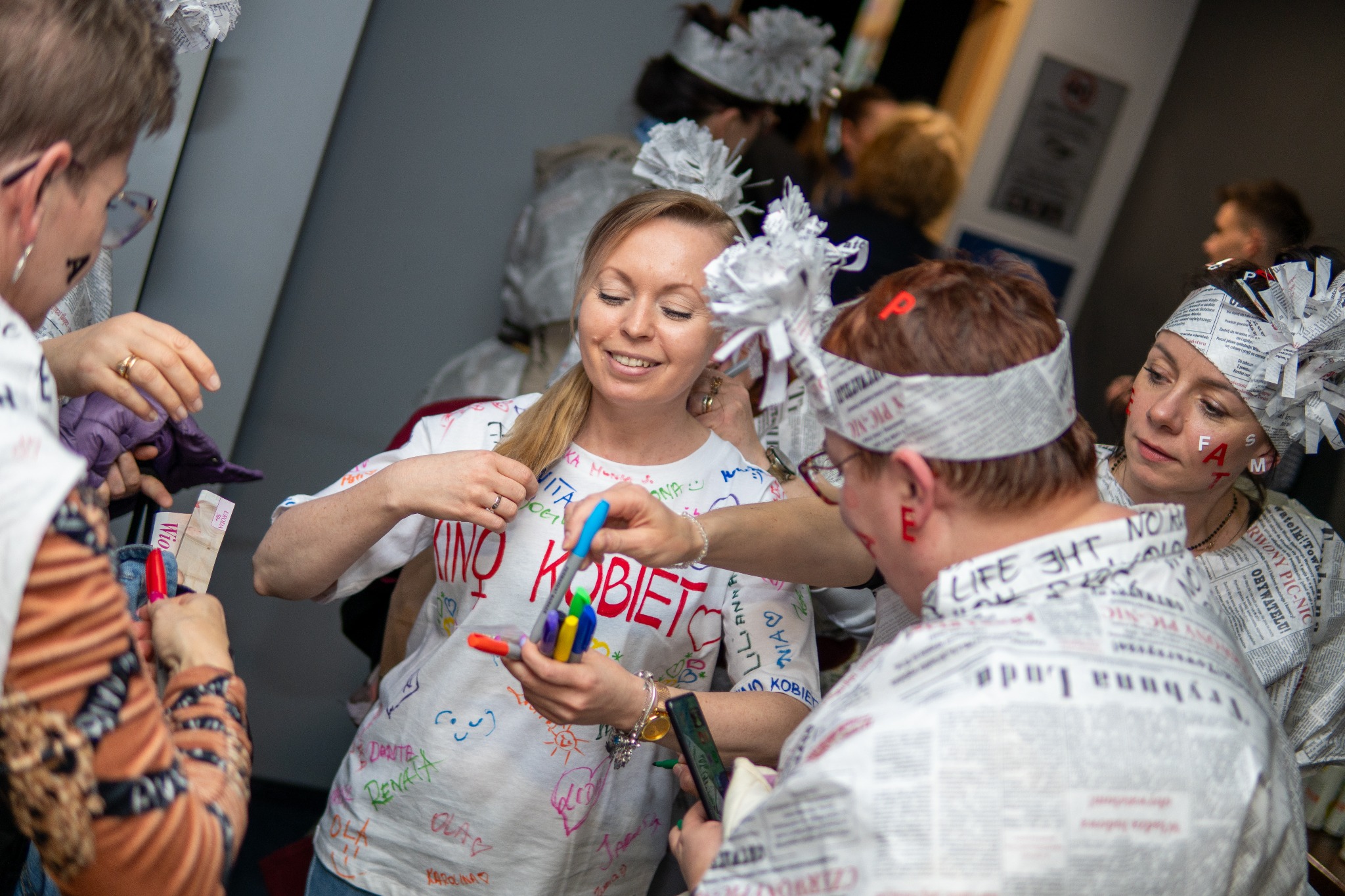 Uśmiechnięta blondynka w tłumie osób w strojach z gazet, trzymająca kolorowe markery, pisząca na koszulce z napisem 'KINO KOBIET'. Nietypowe nakrycia głowy.