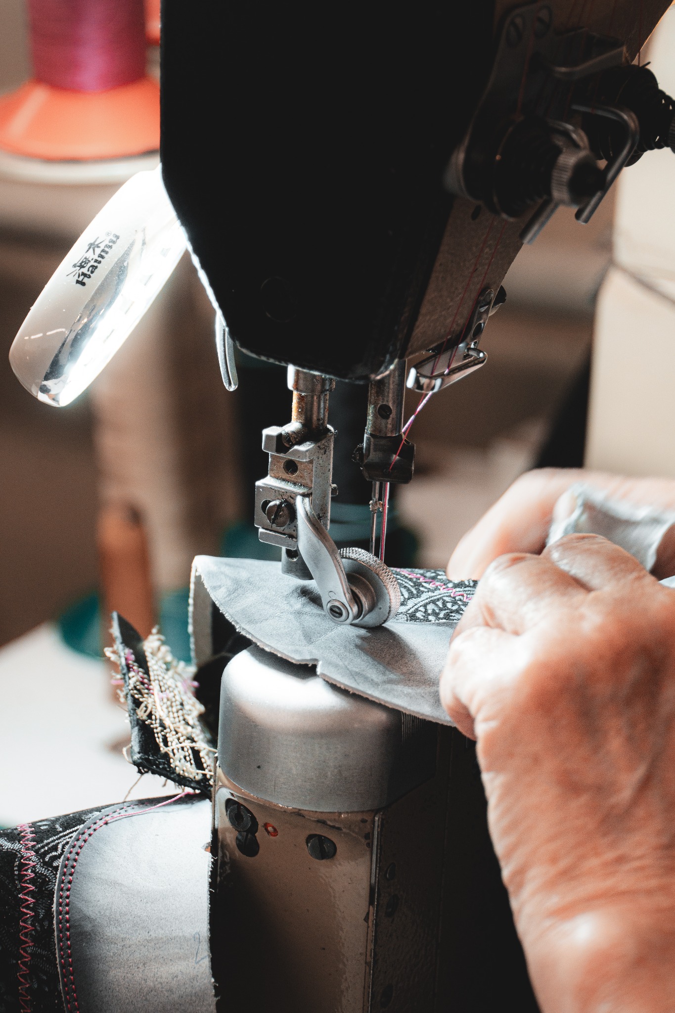 Szczegółowe ujęcie maszyny do szycia w trakcie pracy nad zdobionym materiałem, oświetlonej lampą. Widoczne dłonie operatora i fragment gotowego obszycia.
