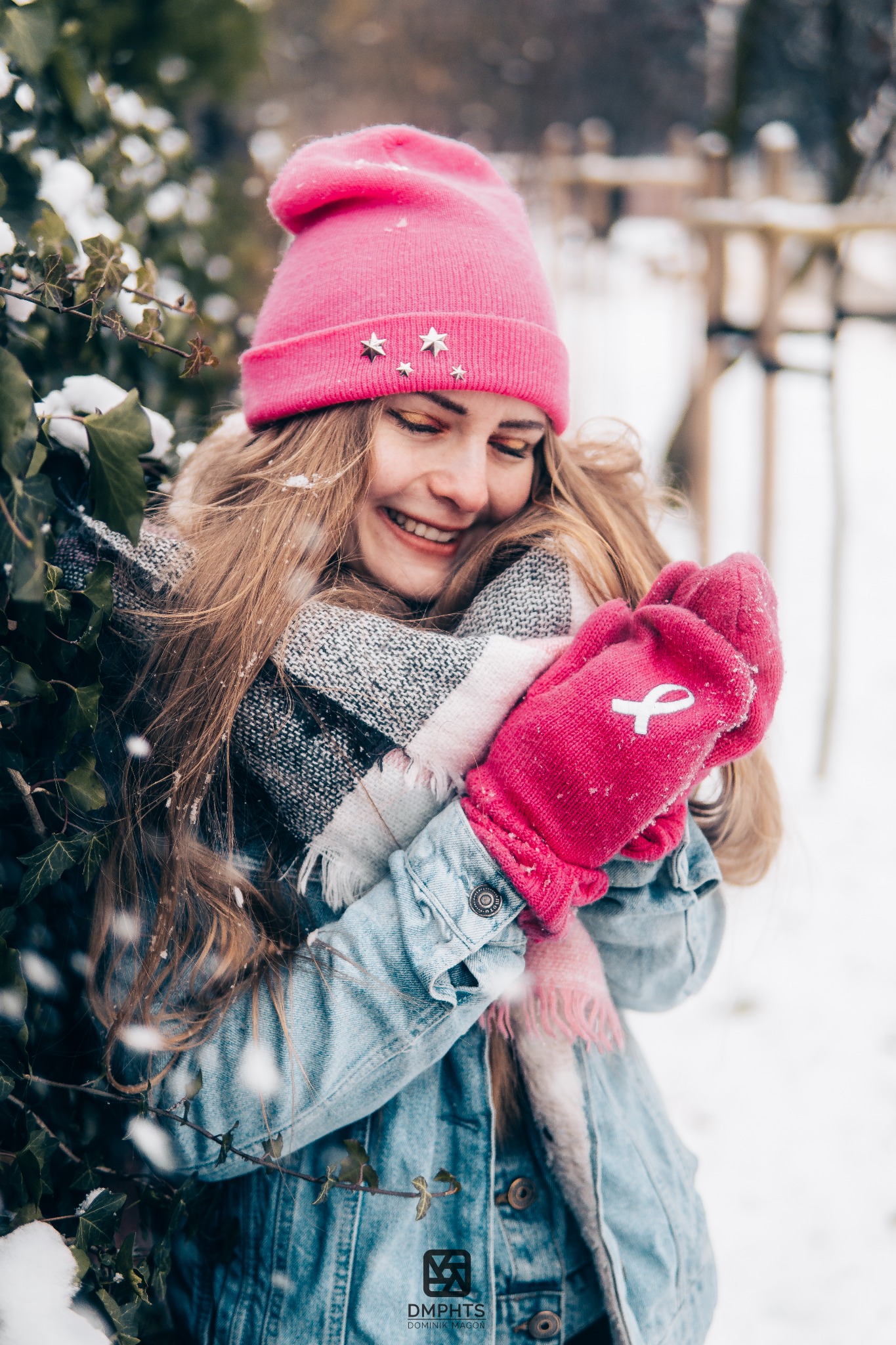 Uśmiechnięta blondynka w różowej czapce i rękawiczkach pozuje w zimowej scenerii w Gnieznie. Śnieg pada, a ona otulona jest szalem i dżinsową kurtką.