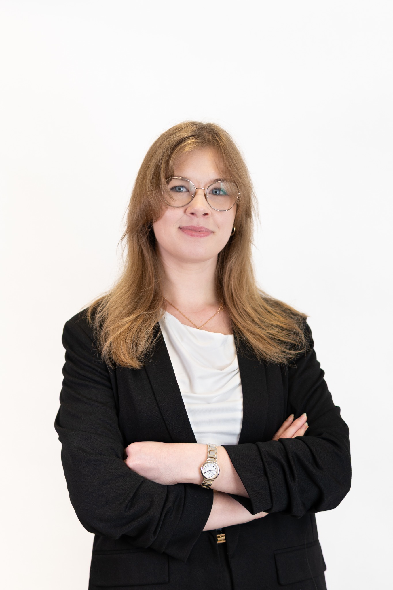 Portret kobiety w okularach, ubrana w czarną marynarkę i białą bluzkę, z założonymi rękami. Jasne tło, naturalne światło, widoczny zegarek na ręce.