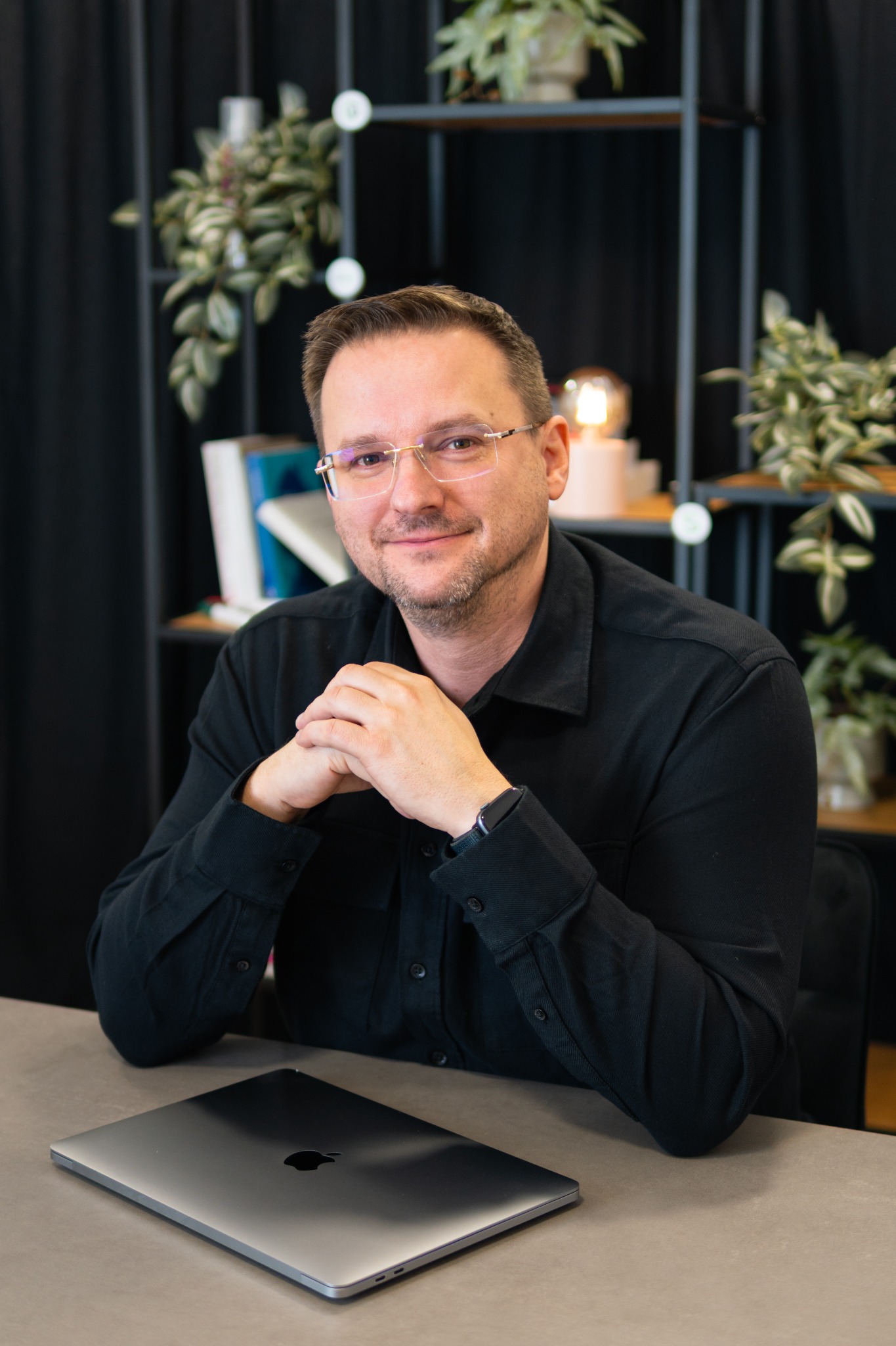Mężczyzna w okularach, w czarnej koszuli, siedzi za biurkiem z laptopem. W tle regał z roślinami i książkami. Uśmiechnięty, z rękami złożonymi na blacie.