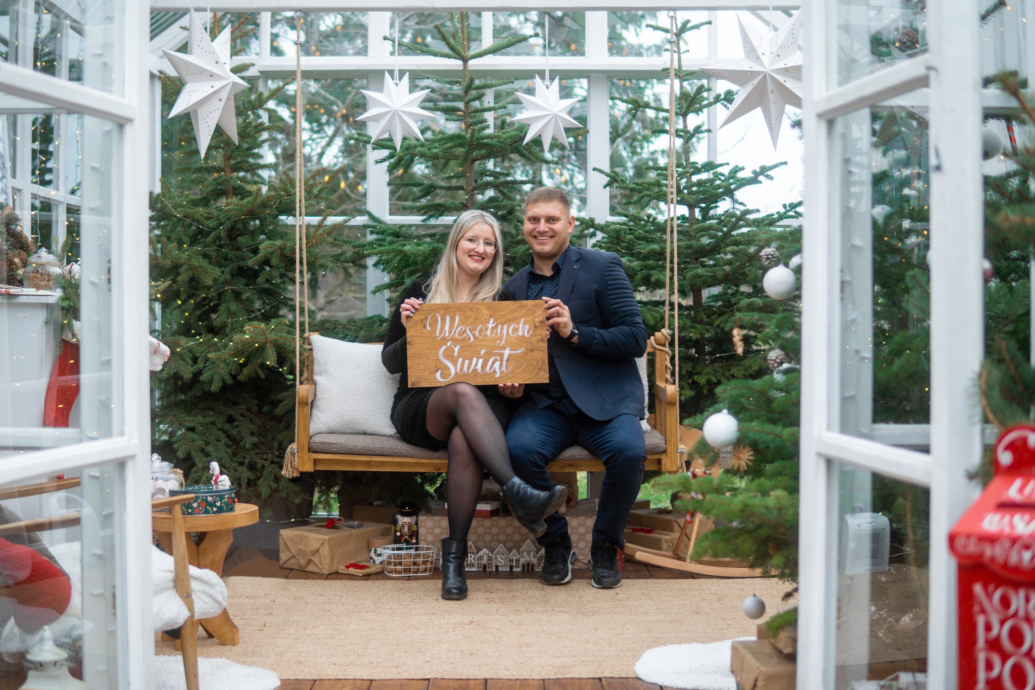 Uśmiechnięta para siedzi na huśtawce w szklarni, trzymając tabliczkę z napisem 'Wesołych Świąt', otoczona choinkami i dekoracjami świątecznymi.