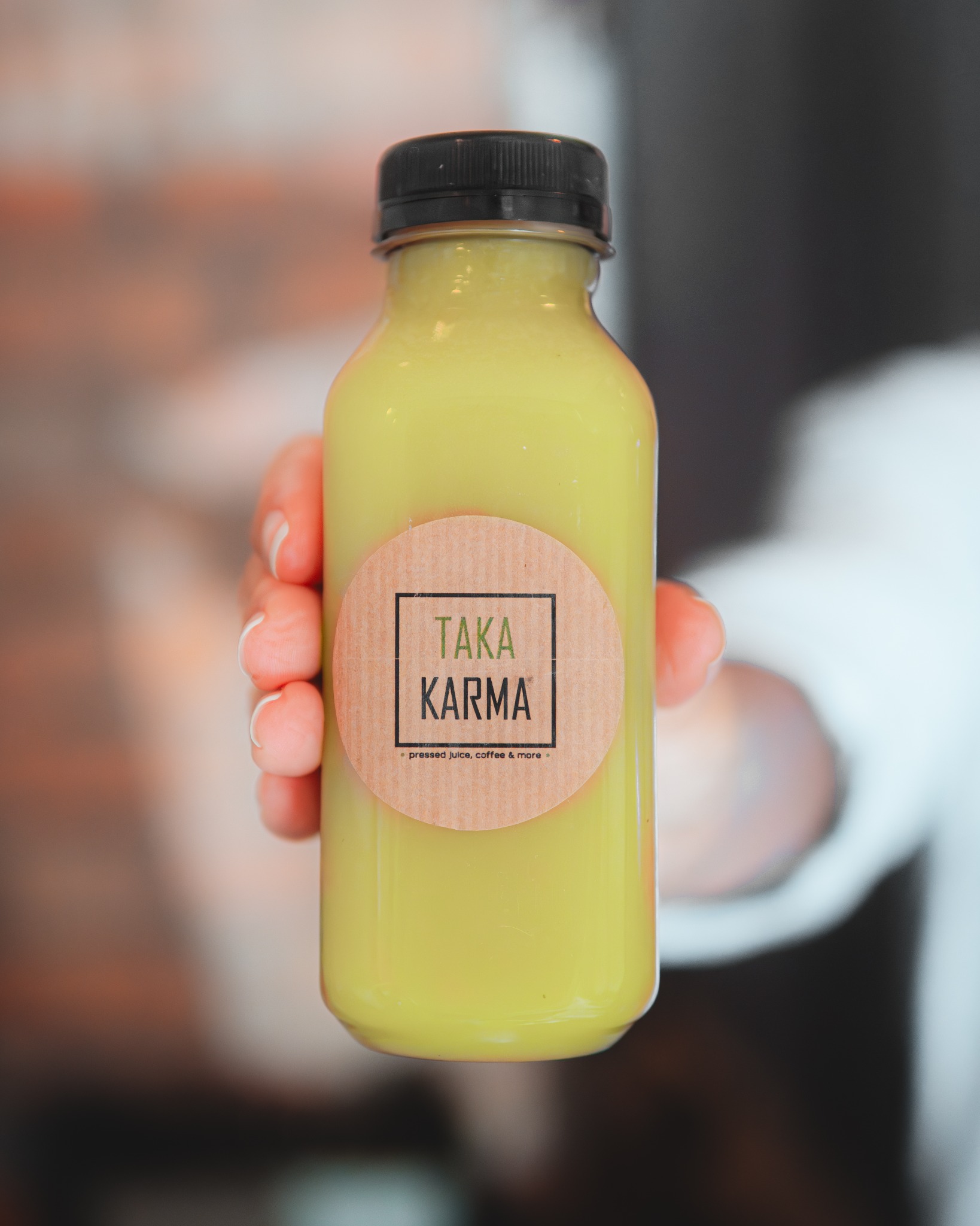Butelka z zielonym sokiem marki Taka Karma trzymana w dłoni, z widocznym logo na etykiecie. Tło rozmyte, z elementami ceglanej ściany i białej koszuli.