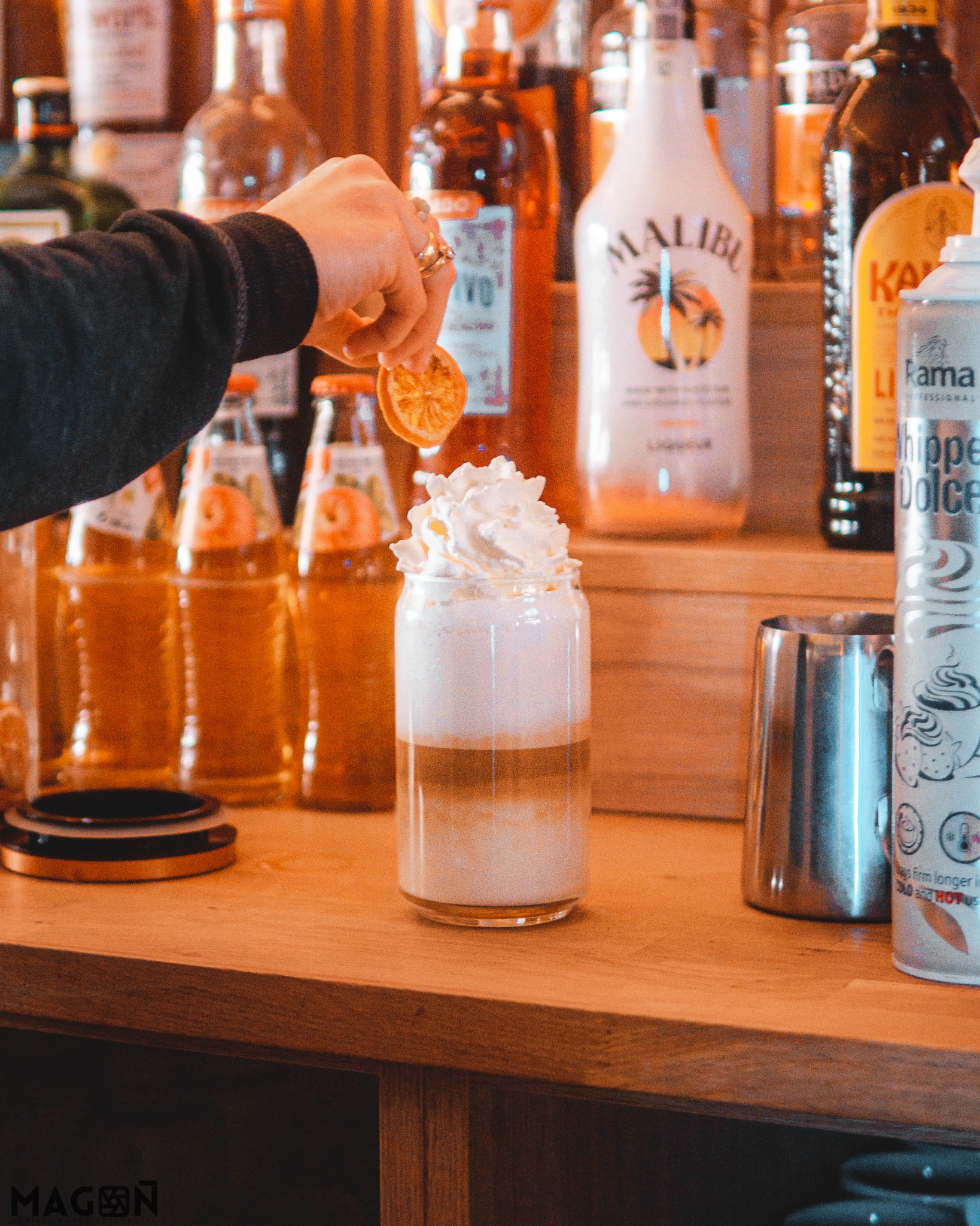 Przygotowywanie warstwowego napoju z bitą śmietaną i plasterkiem pomarańczy na drewnianym barze, w tle butelki alkoholi i syropów.