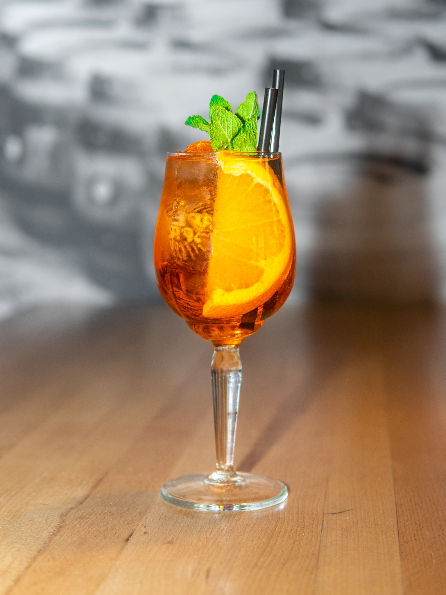 Aperol Spritz w kieliszku na nóżce, z pomarańczą, miętą i słomkami, na drewnianym blacie. Stonowane tło. Fotografia produktowa napoju.