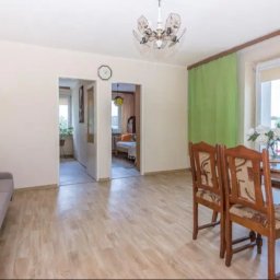 Freedom nieruchomości Białystok Mickiewicza - Jasne wnętrze mieszkania z drewnianą podłogą, sofą z poduszkami i stołem z krzesłami. Widok na pokój przez otwarte drzwi. Lampa sufitowa i okno z zieloną zasłoną.