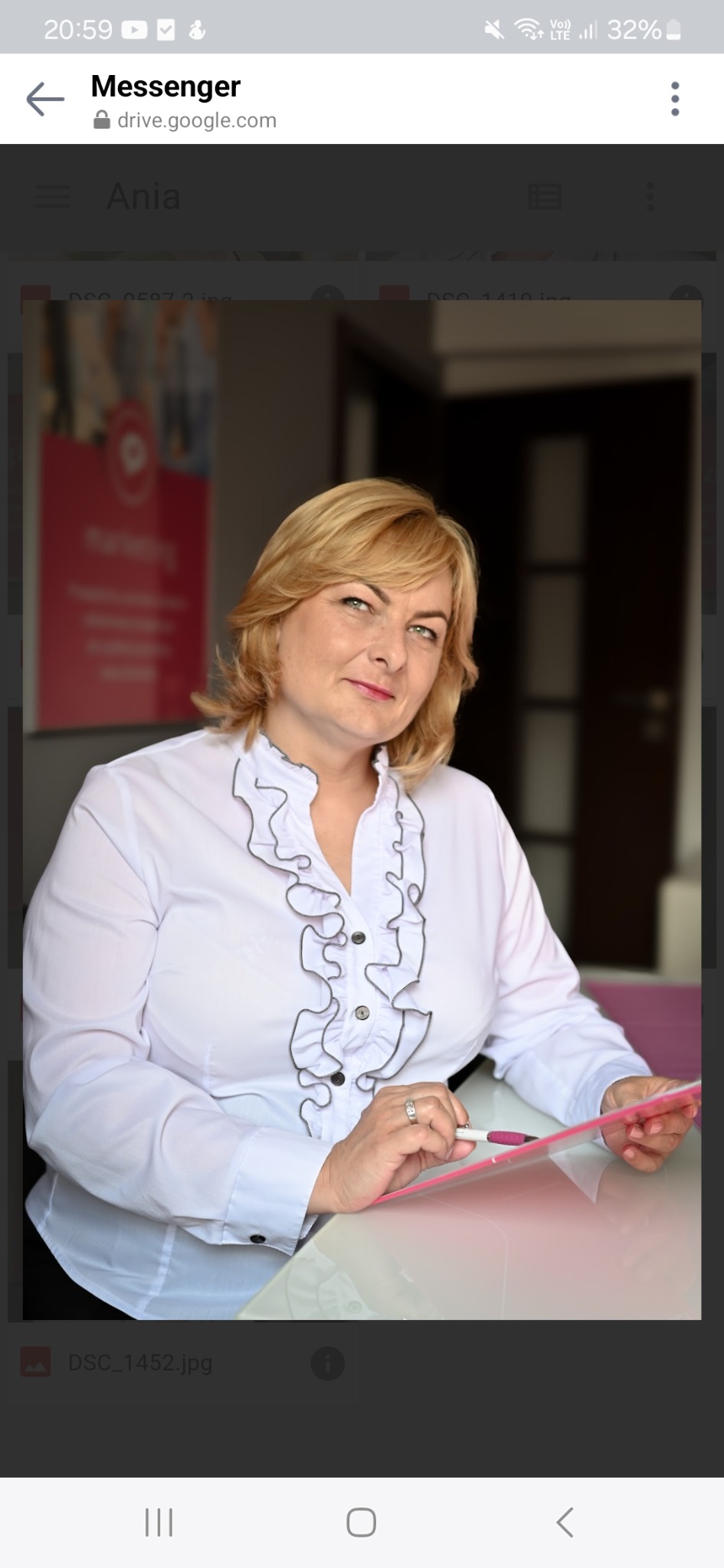 Portret kobiety w białej bluzce z falbanami, trzymającej różową teczkę i długopis. W tle biuro z plakatem marketingowym i ciemnymi drzwiami.