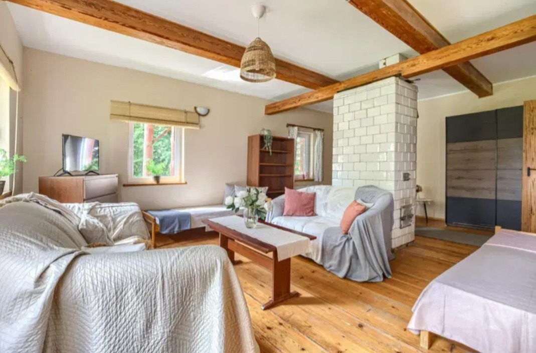 Jasne wnętrze pokoju z drewnianymi belkami stropowymi, białym piecem kaflowym i drewnianą podłogą. Widoczne sofy, łóżka i stół. W tle okno z widokiem na zieleń.