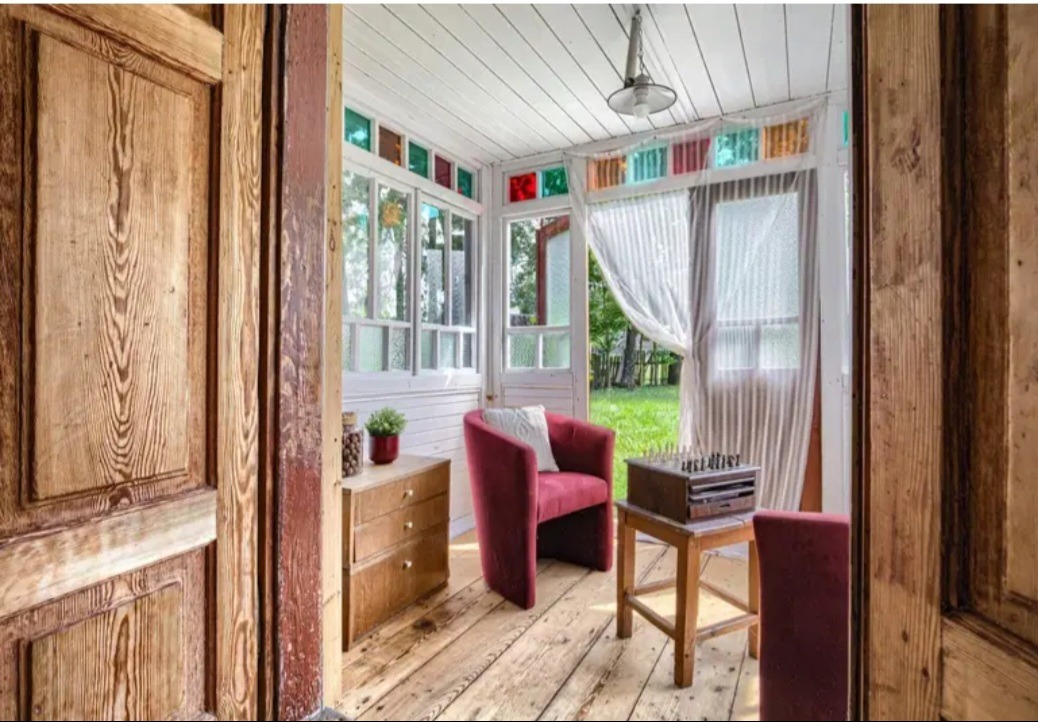 Jasny, przeszklony ganek z drewnianą podłogą i meblami w stylu retro. Widoczne krzesła, stolik z szachami i komoda. Kolorowe szkła w oknach dodają unikalnego charakteru.