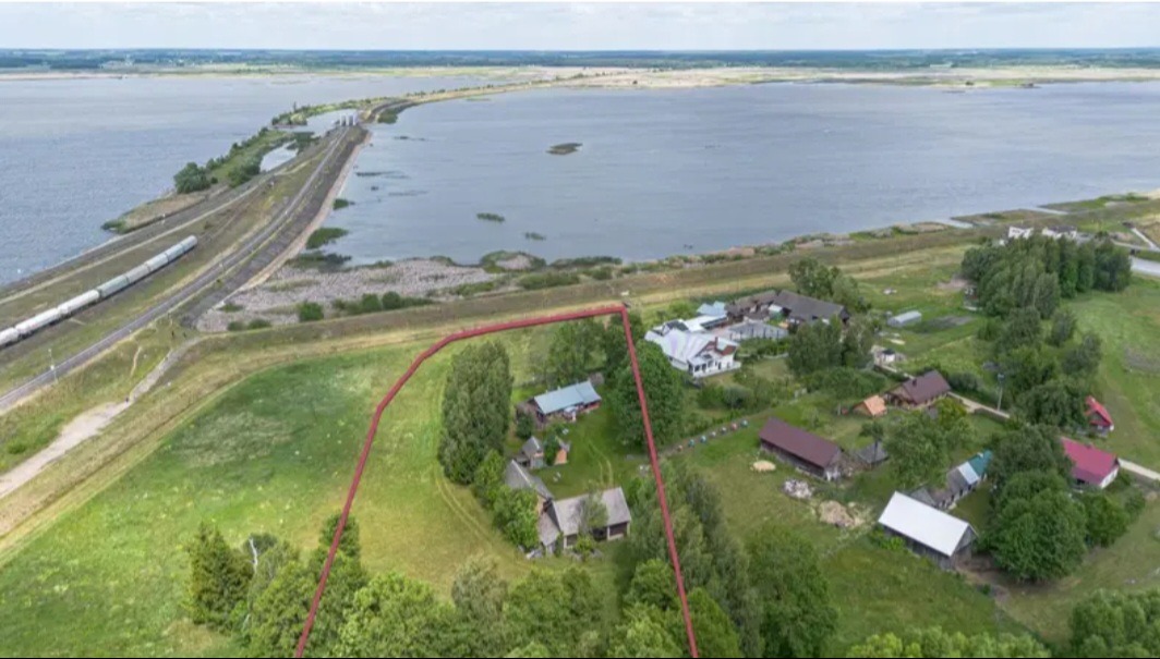 Panoramiczny widok z drona na działkę z domami, otoczoną czerwoną linią, w tle jezioro i linia kolejowa z pociągiem towarowym. Ujęcie z góry, słoneczny dzień.