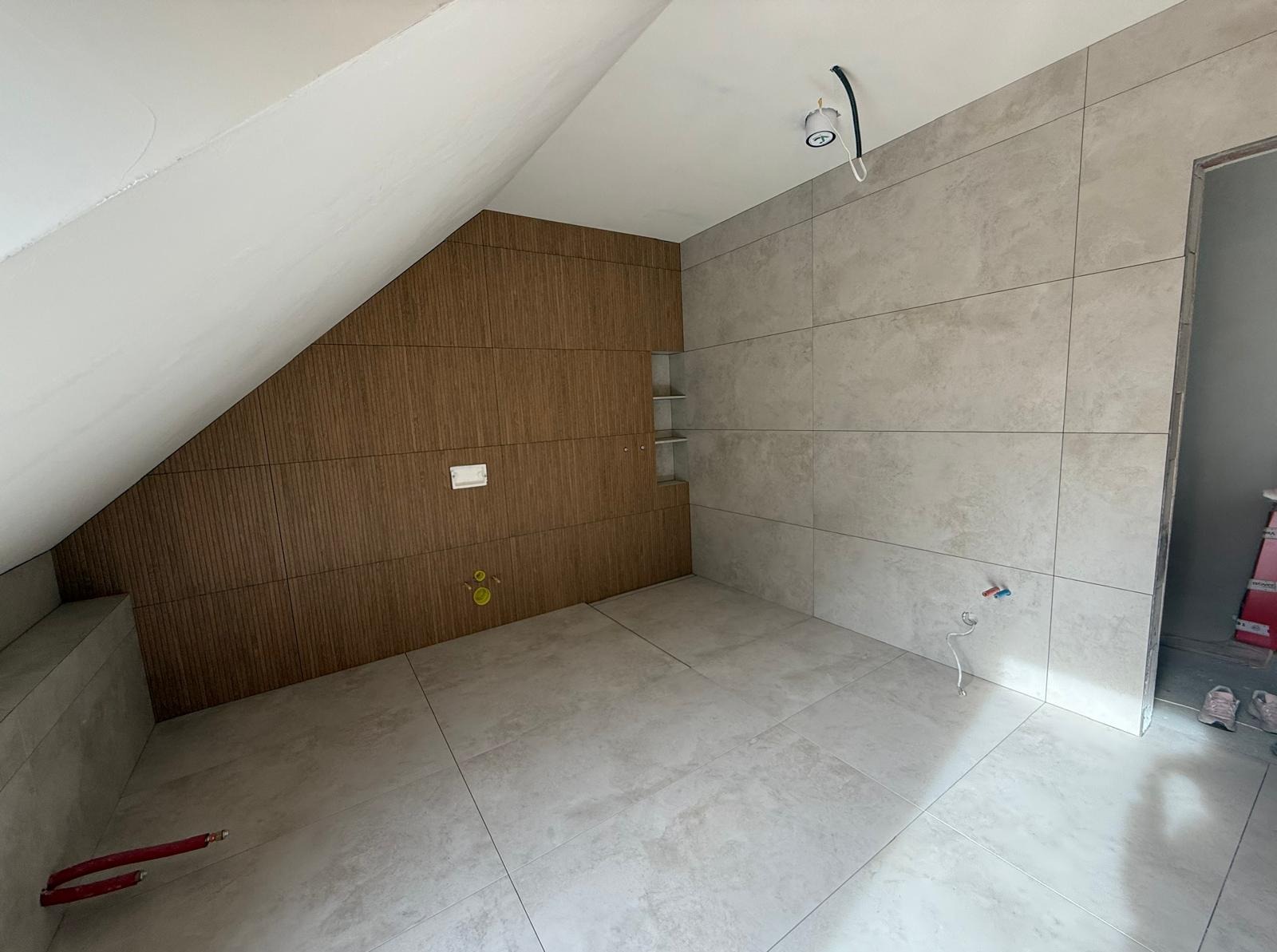 Wykończona łazienka na poddaszu z brązową ścianą z drewnopodobnych paneli i szarą ścianą z płytek. Widoczne instalacje hydrauliczne i oświetlenie.