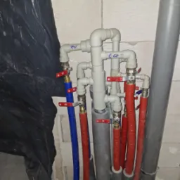 Instalacja hydrauliczna z rurami w kolorze niebieskim i czerwonym, z zaworami i izolacją, na tle ściany z betonu kom&oacute;rkowego i czarnej folii.