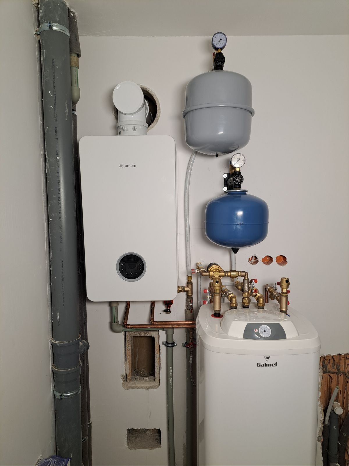 Biały piec gazowy Bosch i zbiornik Galmet z mosiężnymi rurami w kotłowni. Szare rury odprowadzające spaliny. Manometry na zbiornikach. Widoczne ślady po kuciu.