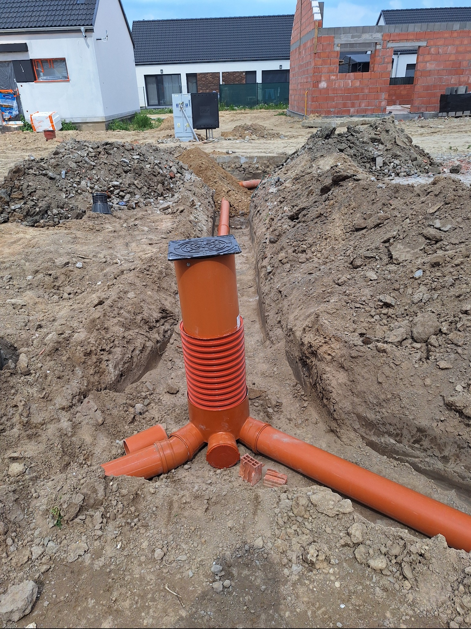 Pomarańczowa studzienka kanalizacyjna z pokrywą, w wykopie ziemnym na tle budynków w trakcie budowy. Widoczne rury kanalizacyjne i gruz.