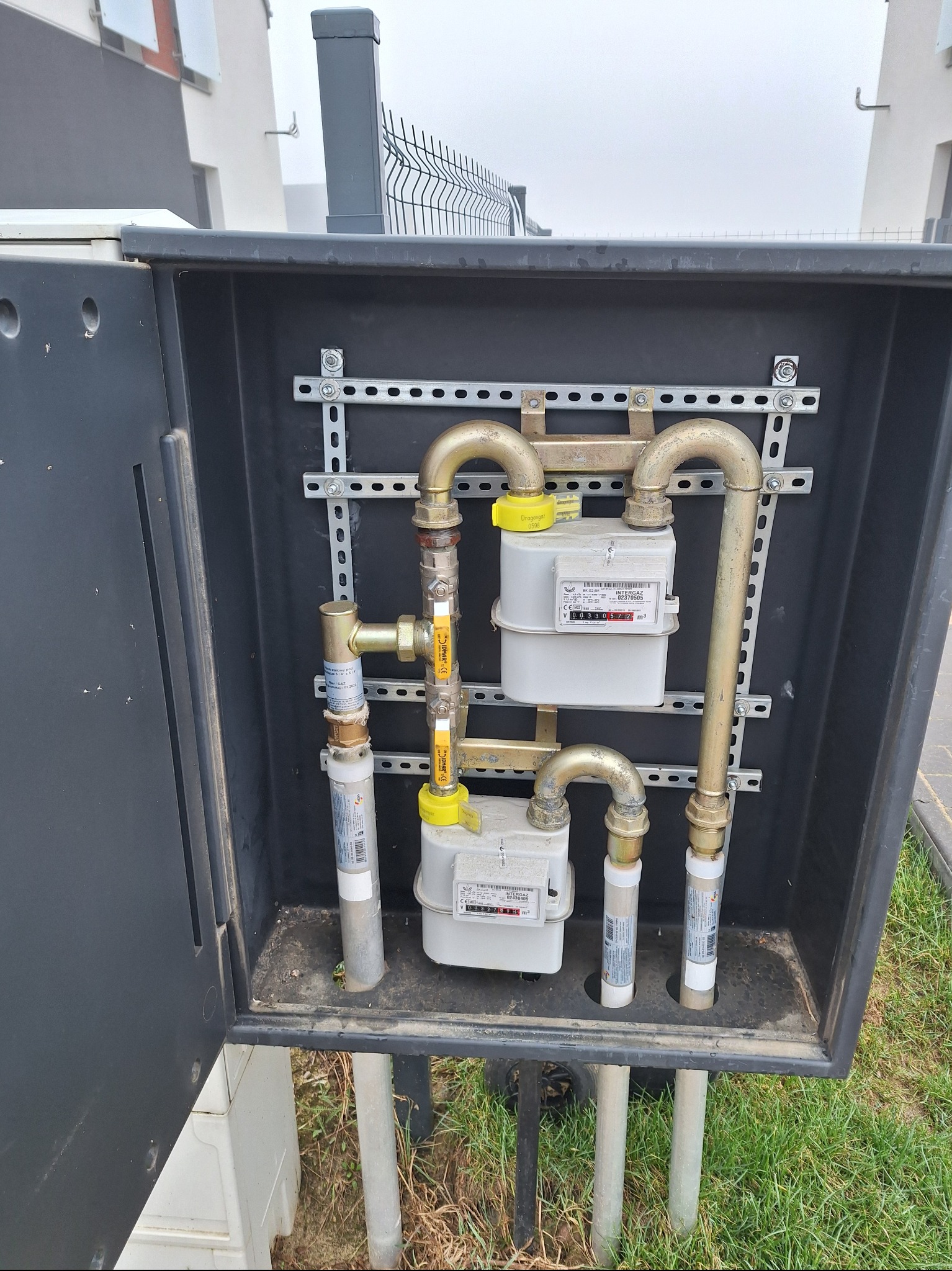 Dwa liczniki gazu INTERGAZ w czarnej skrzynce, zamontowane na metalowych szynach. Widoczne rury gazowe z żółtymi oznaczeniami. Na zewnątrz skrzynki trawa i fragment ogrodzenia.