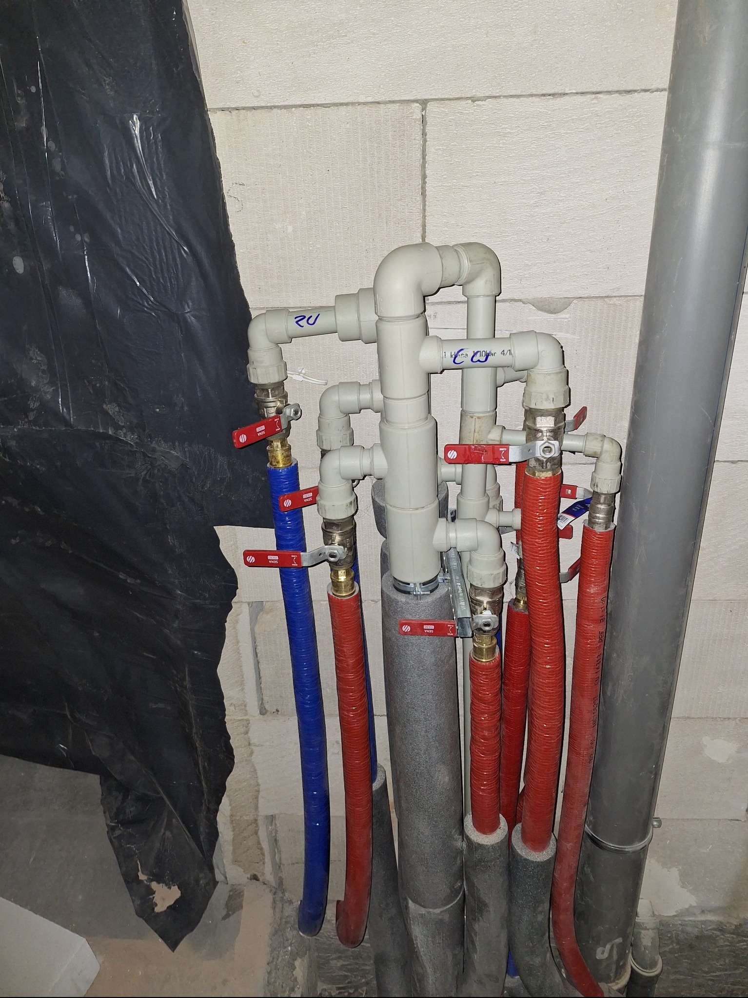 Instalacja hydrauliczna z rurami w kolorze niebieskim i czerwonym, z zaworami i izolacją, na tle ściany z betonu komórkowego i czarnej folii.